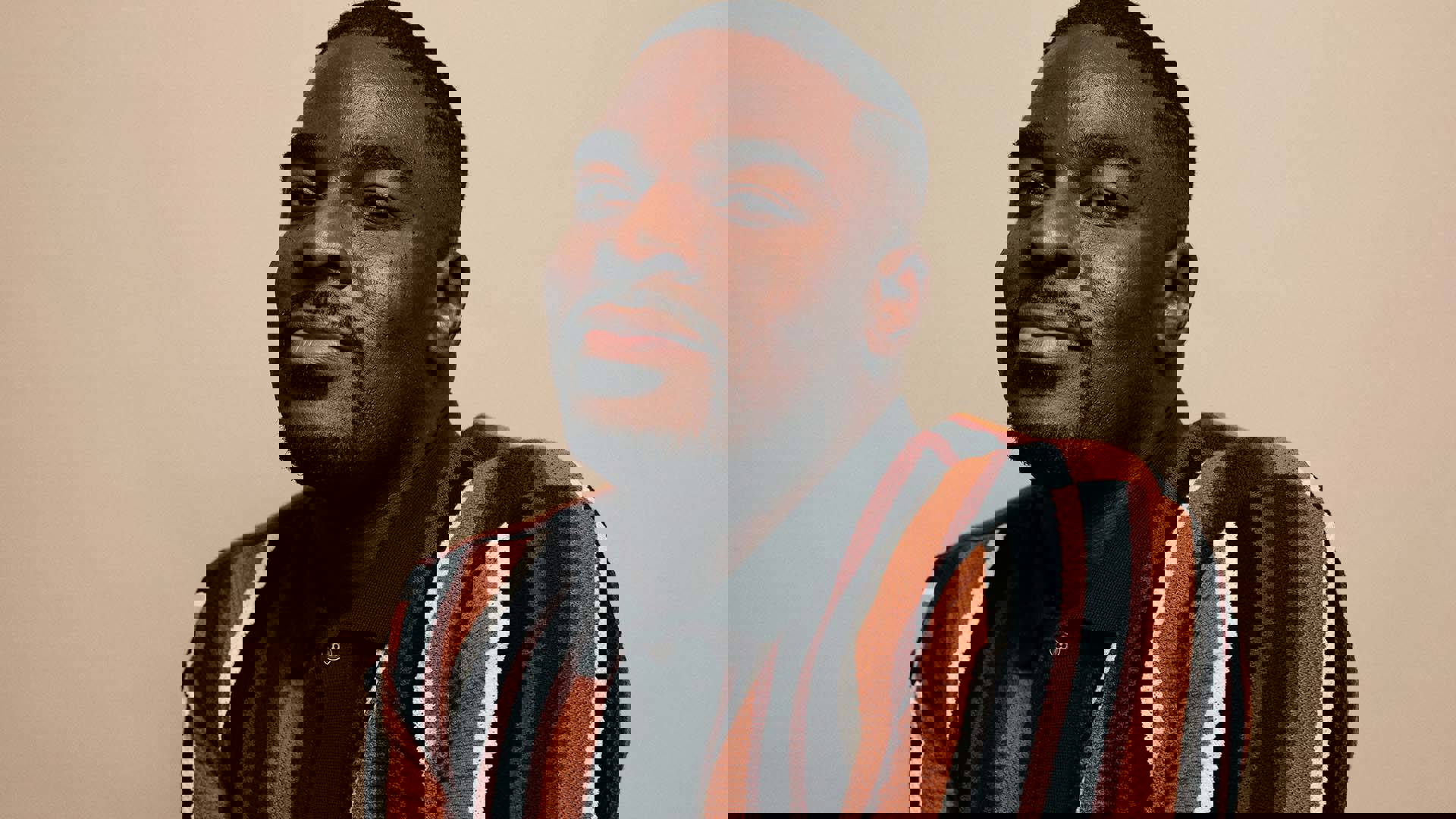 Kyle Taylor Parker, a black man with dark complexion, a goatee and short, buzzed black hair. He is looking at the camera with a slight smile, wearing an orange, brown and white striped knit polo against a tan background.