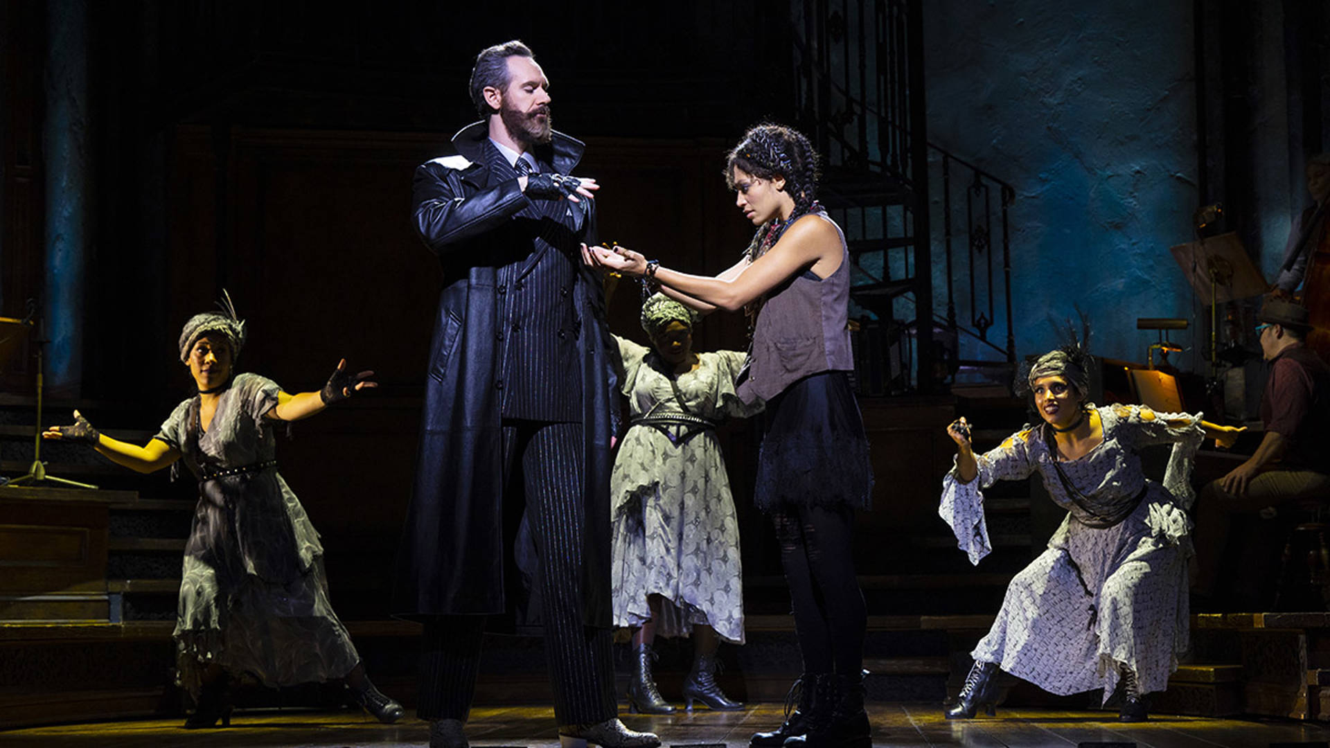 A tall caucasian man in a trench coat with a brown beard is crossing his arms. A younger woman nearby holds her hands out to him.