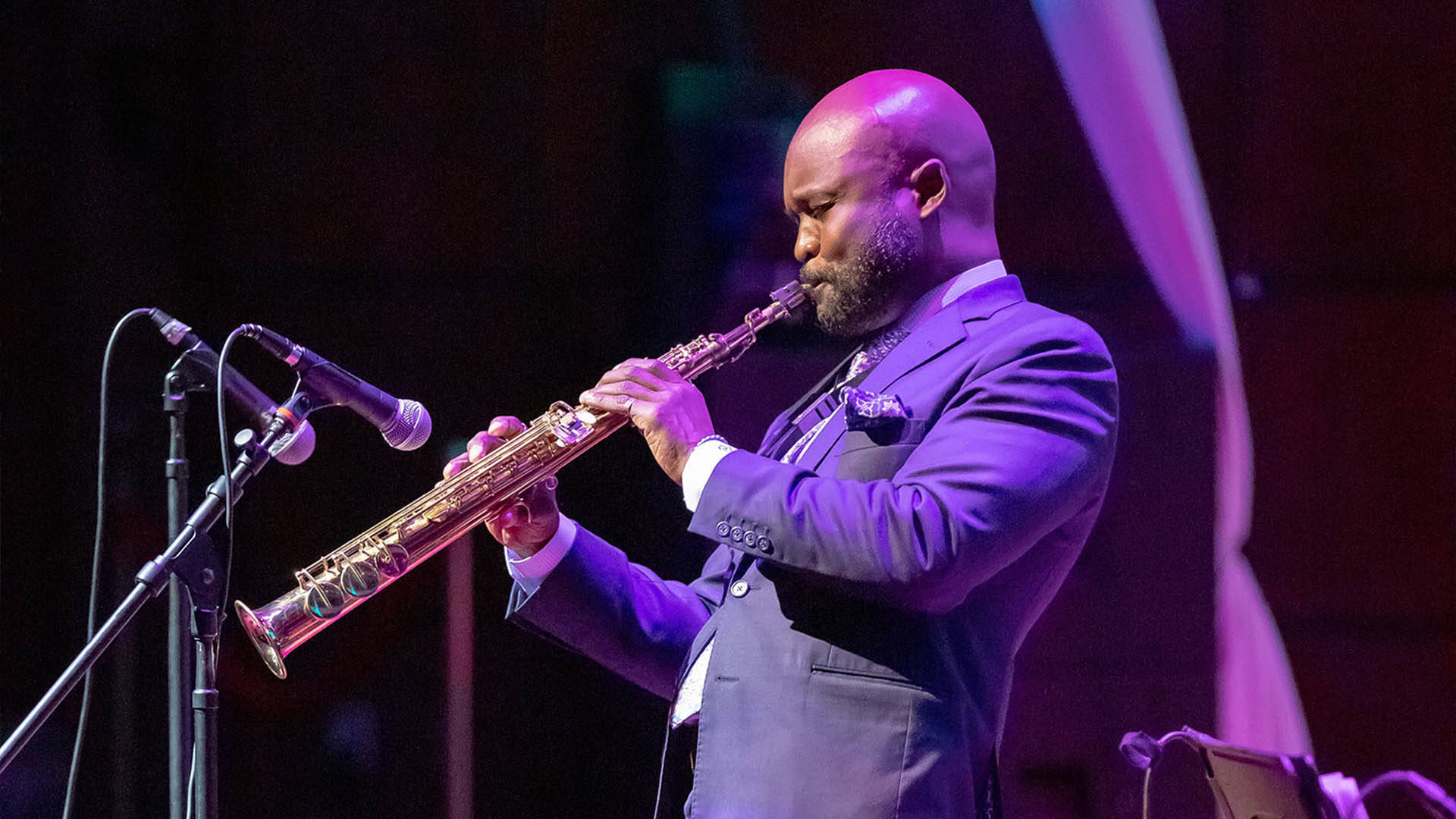 A bald, Black man with a short, dark beard playing an alto saxophone. He is wearing a blue suit.