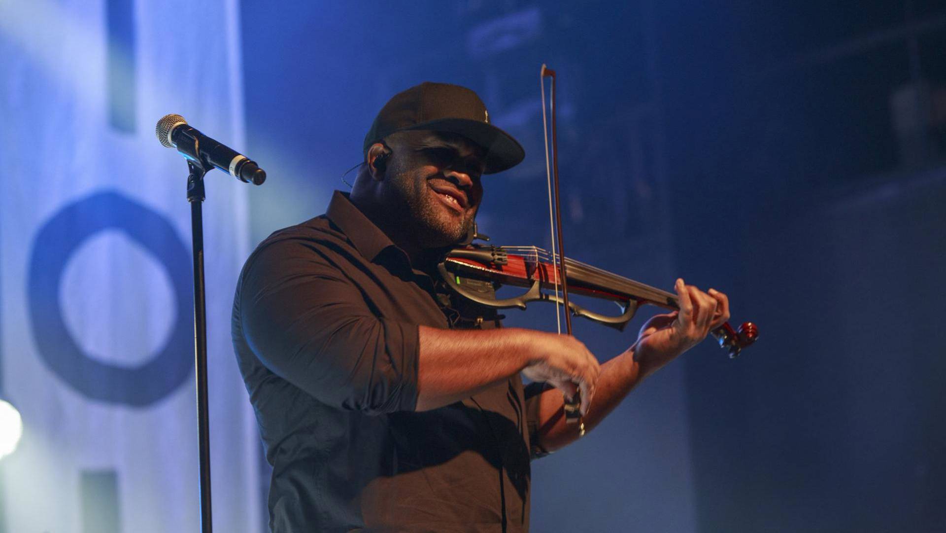 A man with dark complexion, a dark shirt, and a dark baseball cap plays an electric violin.