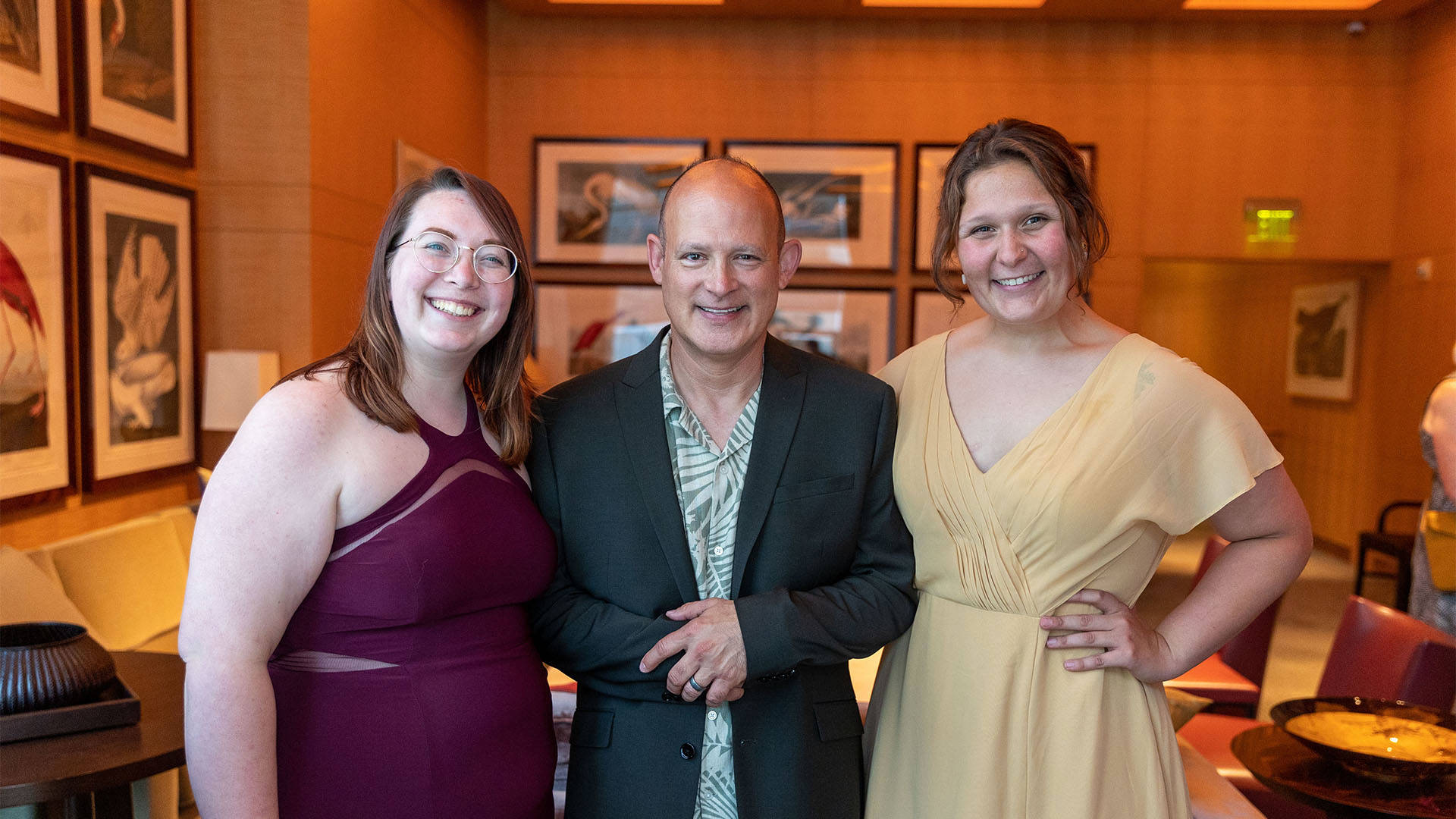Two young caucasian women with brown hair and dresses stand with an older man with a bald head, jacket and floral shirt.