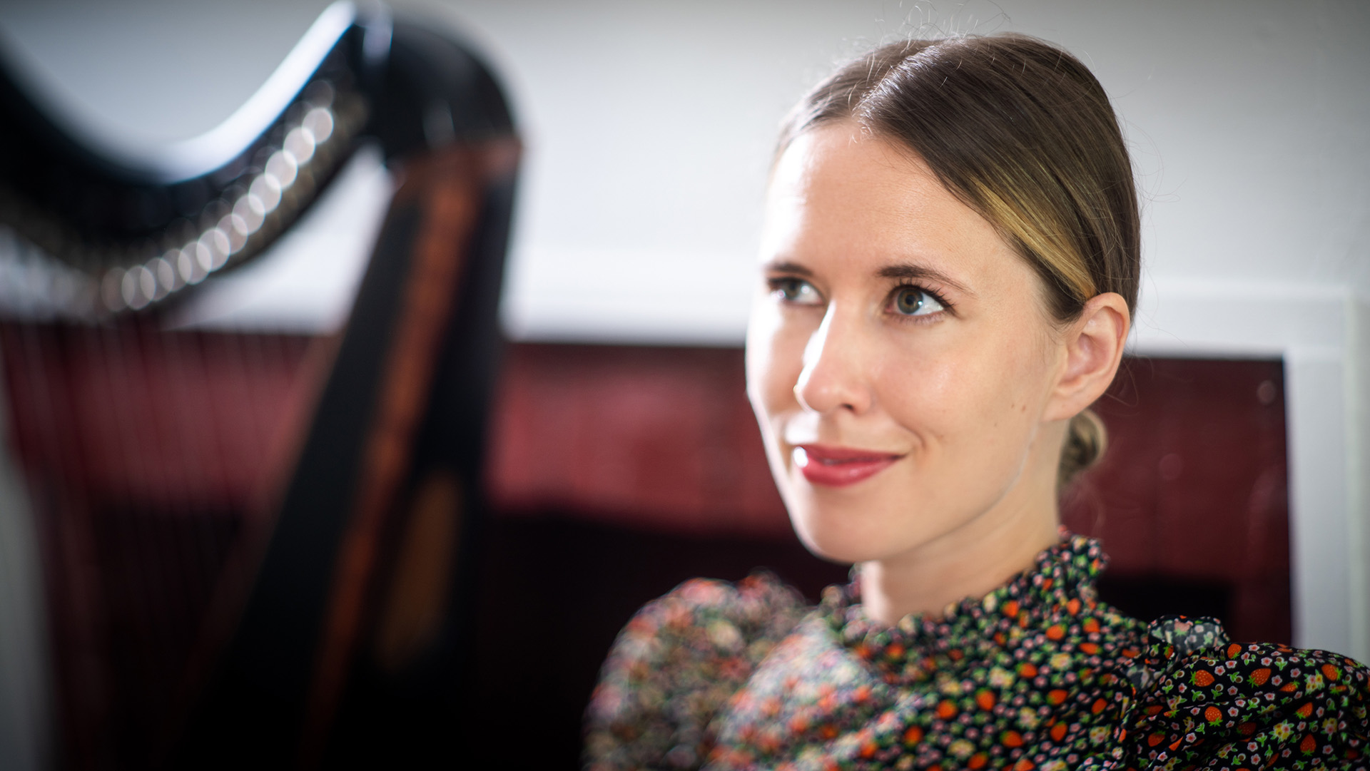 A caucasian woman with dark hair pulled back and brown eyes is smiling. In the background there is a blurry harp. She has a colorful top on.