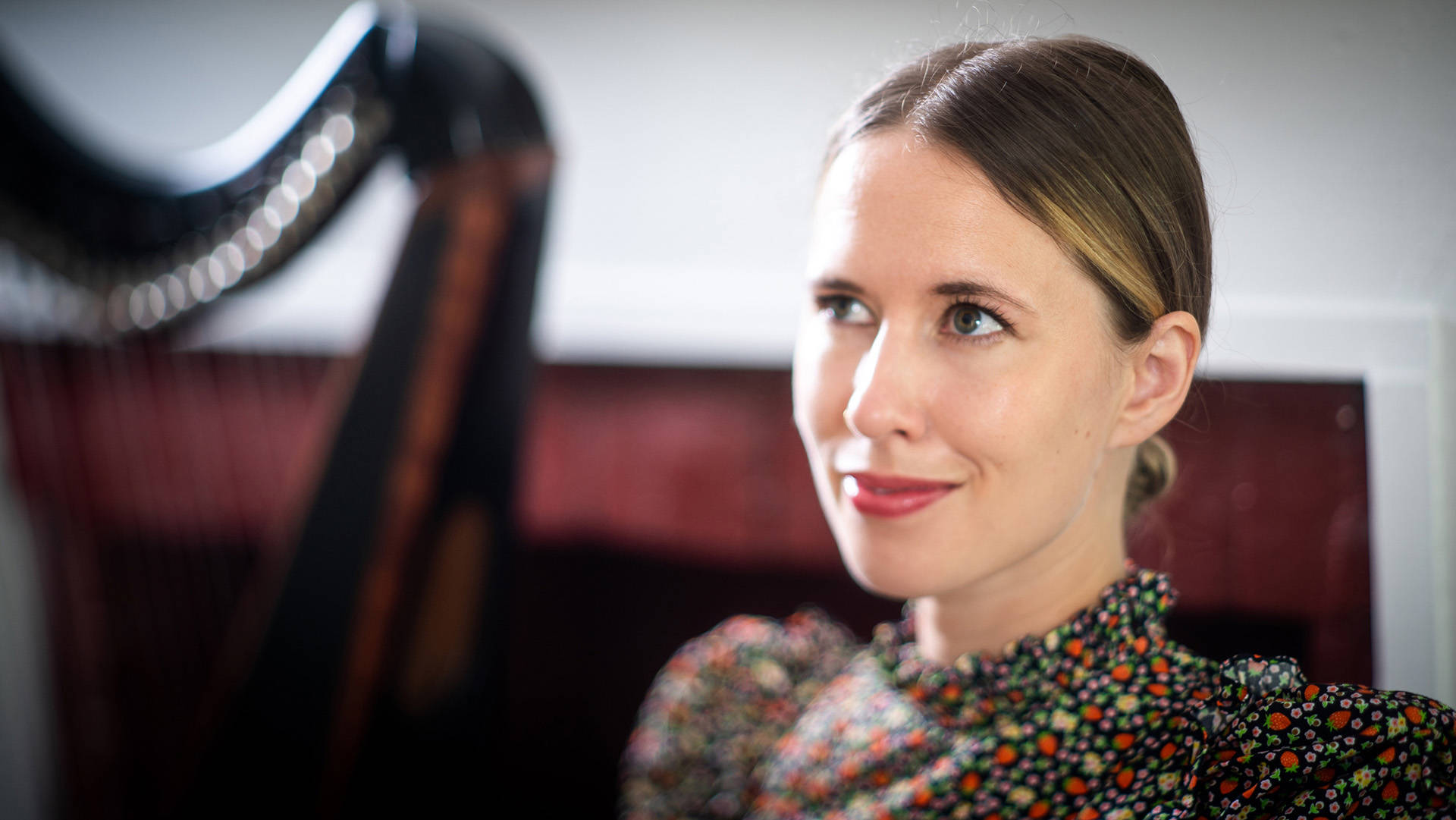 A caucasian woman with dark hair pulled back and brown eyes is smiling. In the background there is a blurry harp. She has a colorful top on.