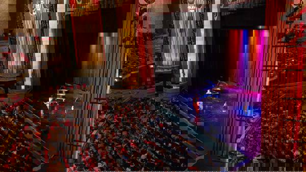 A wide shot of Capitol Theater and an audience of young students. On stage a person is making announcements in front of a piano and a guitar.