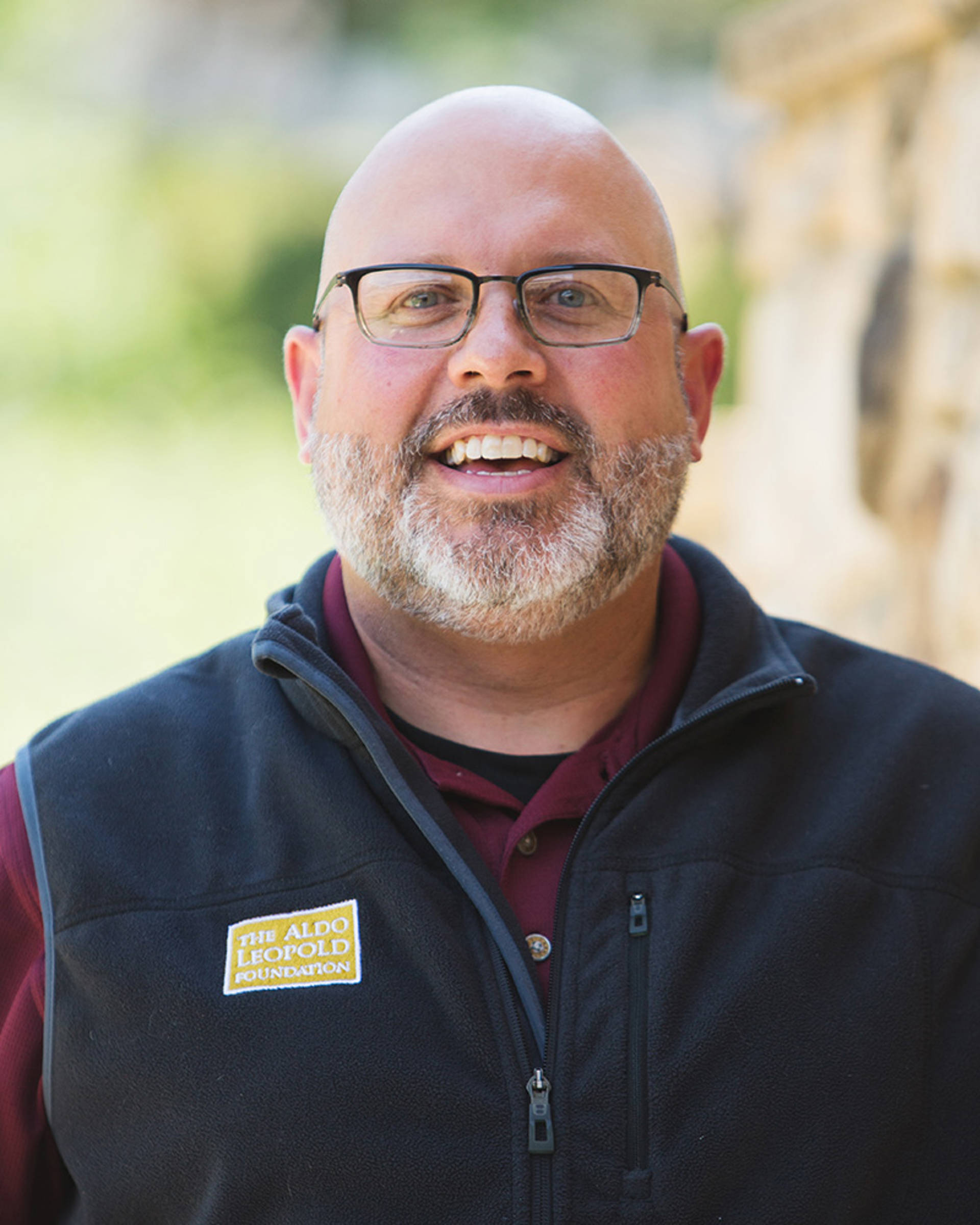 Headshot of a middleaged smiling man with light complexion, a bald head, short grey beard, and glasses.