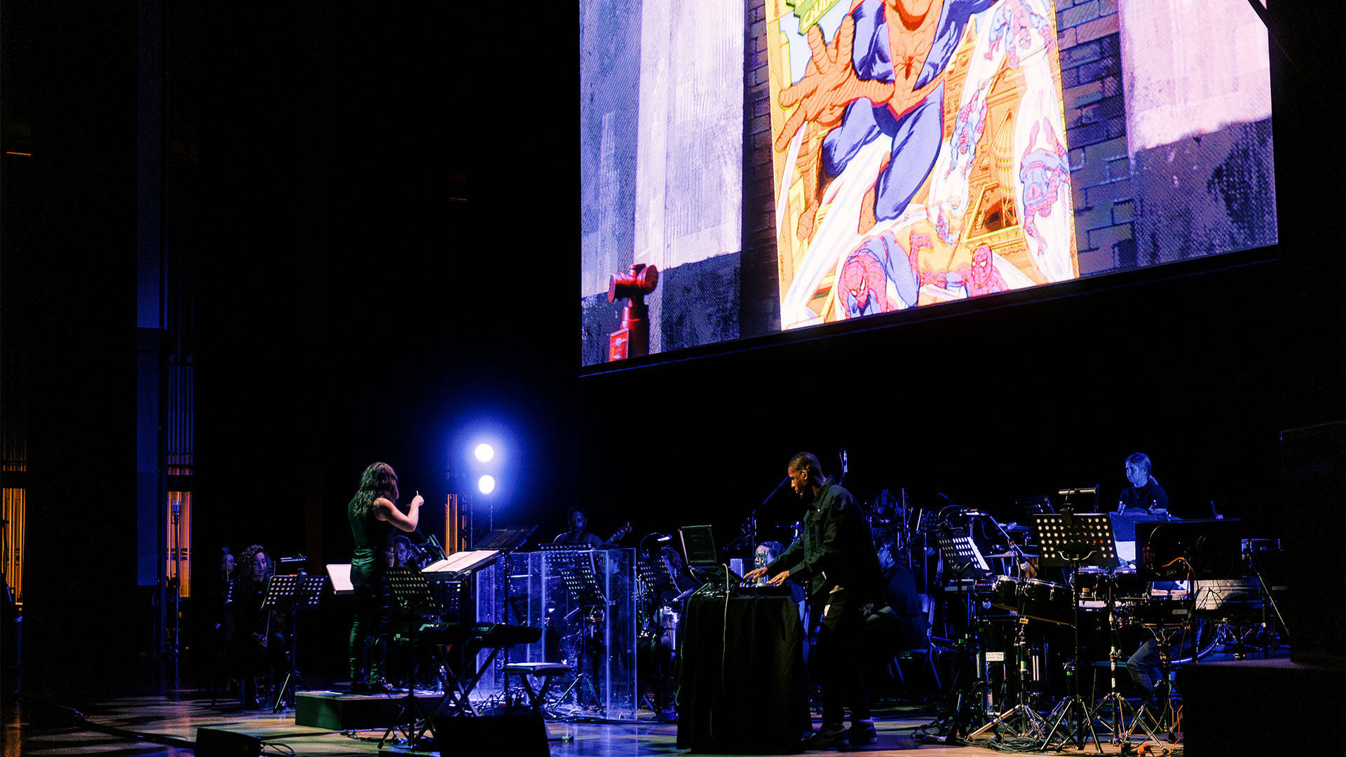 A woman with light complexion and long dark hair in a dark outfit conducting an orchestra. A screen above them is playing a scene from Spiderman: Into the Spiderverse.
