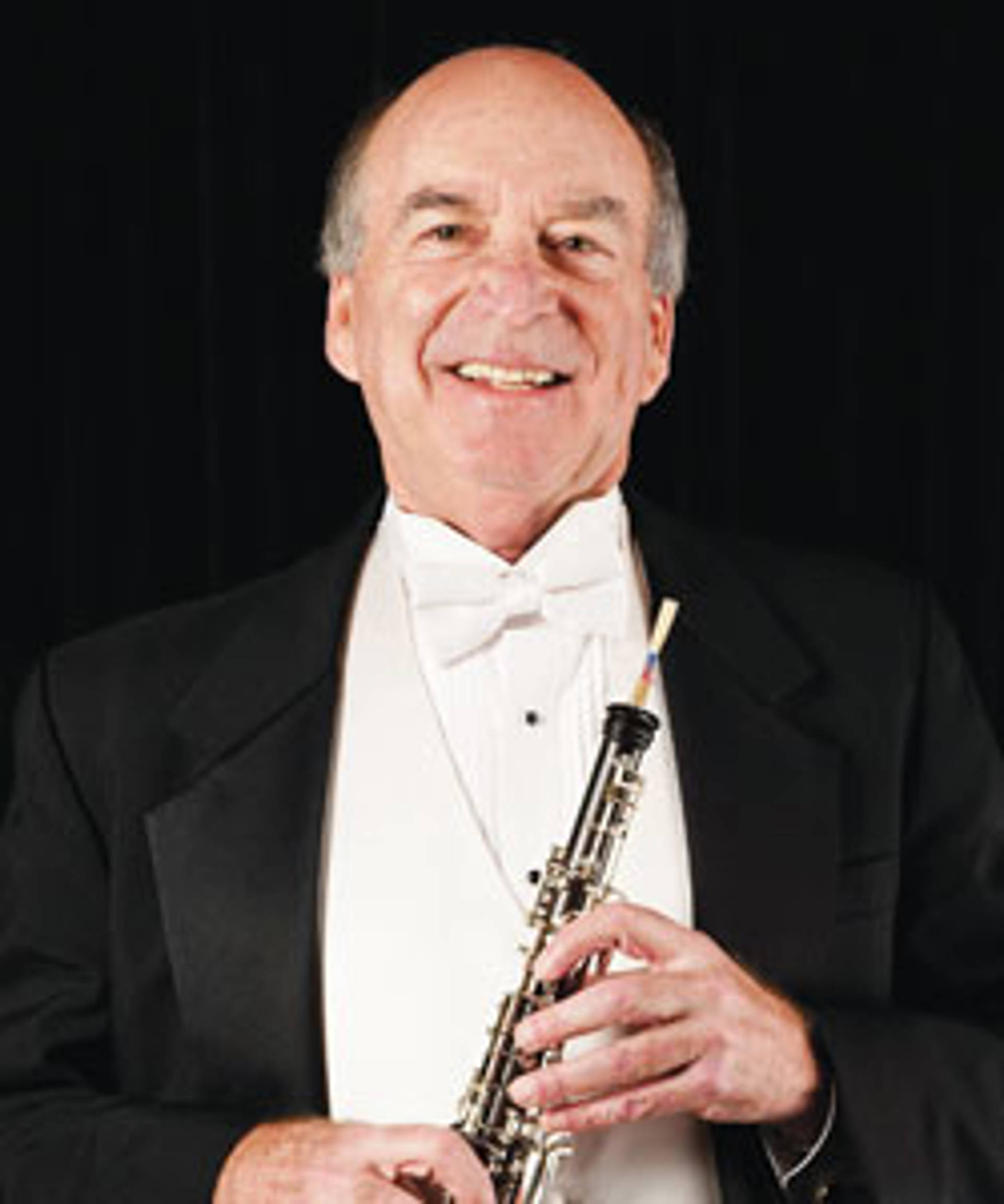 Headshot of a caucasian man holding an oboe in a tux. He has a bald head with grey hair on the sides