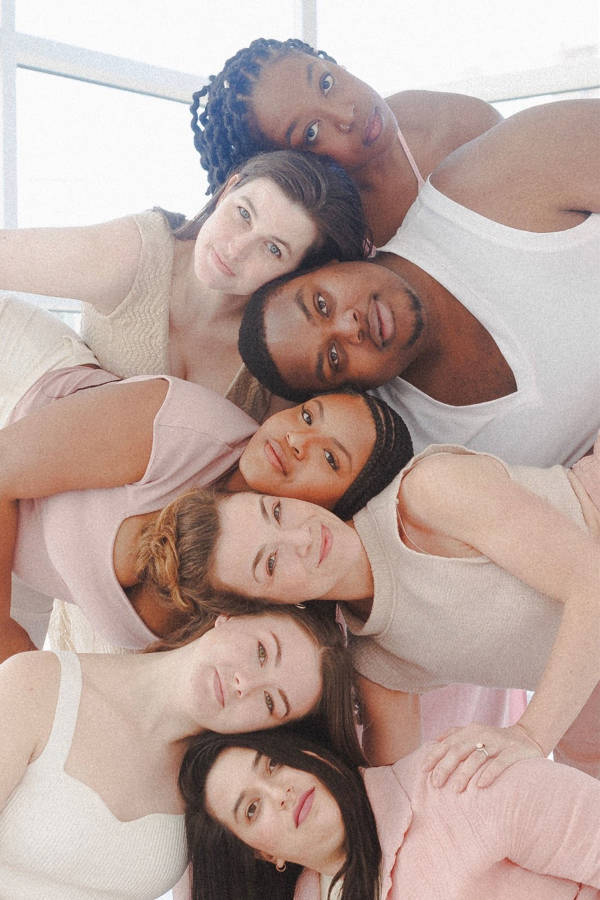 Seven dancers in flowing white, beige and pink outfits leaning in and stacking their heads for a photo.
