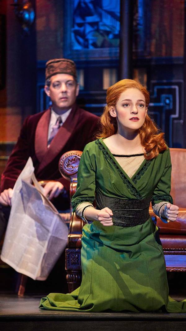 A women with red hair and a green dress looking off in the distance. A man in a red coat sits nearby holding a newspaper