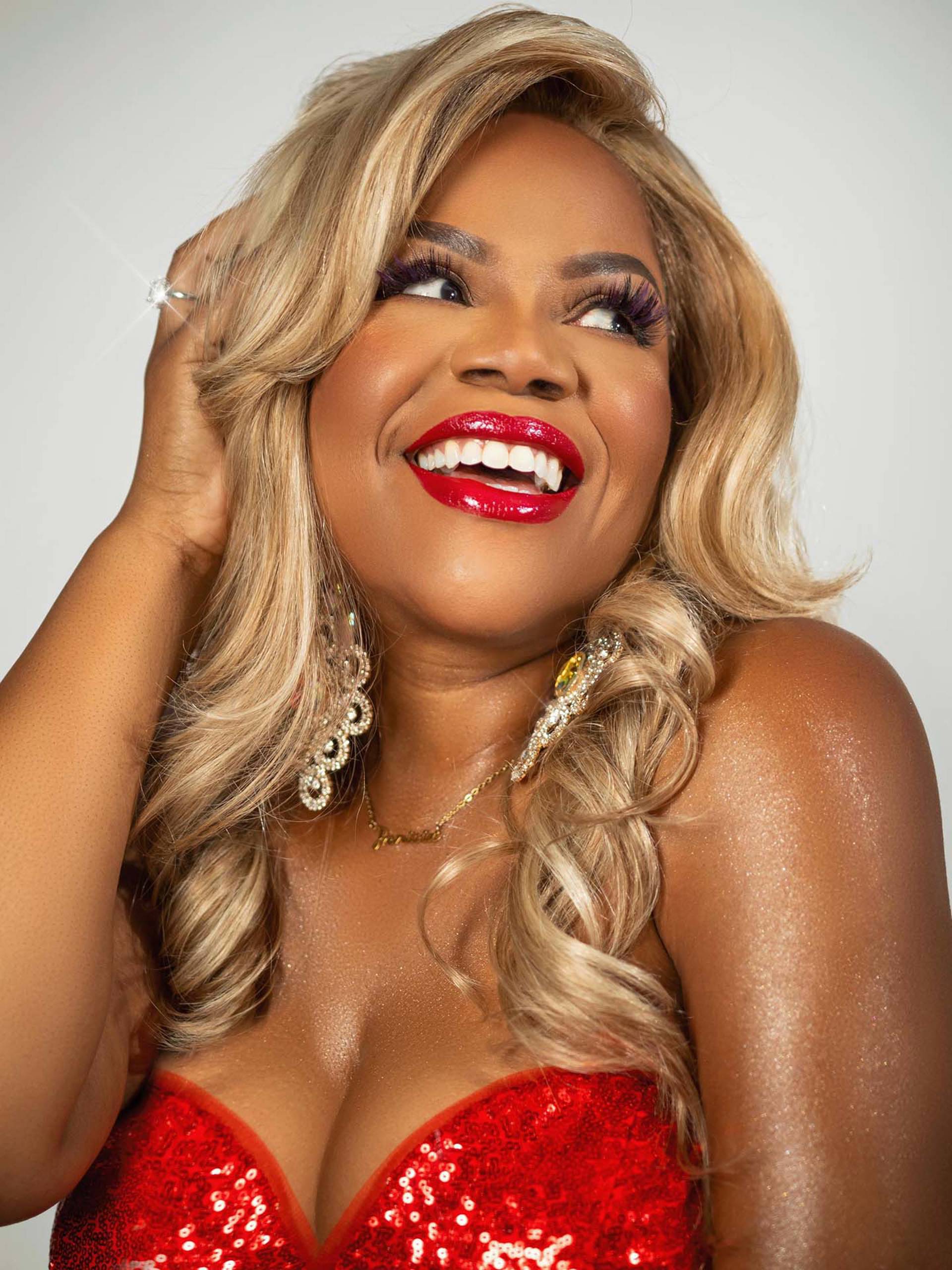 A headshot of a Black woman with brown eyes and blonde hair in a red, sequined top. She has on bright, red lipstick.