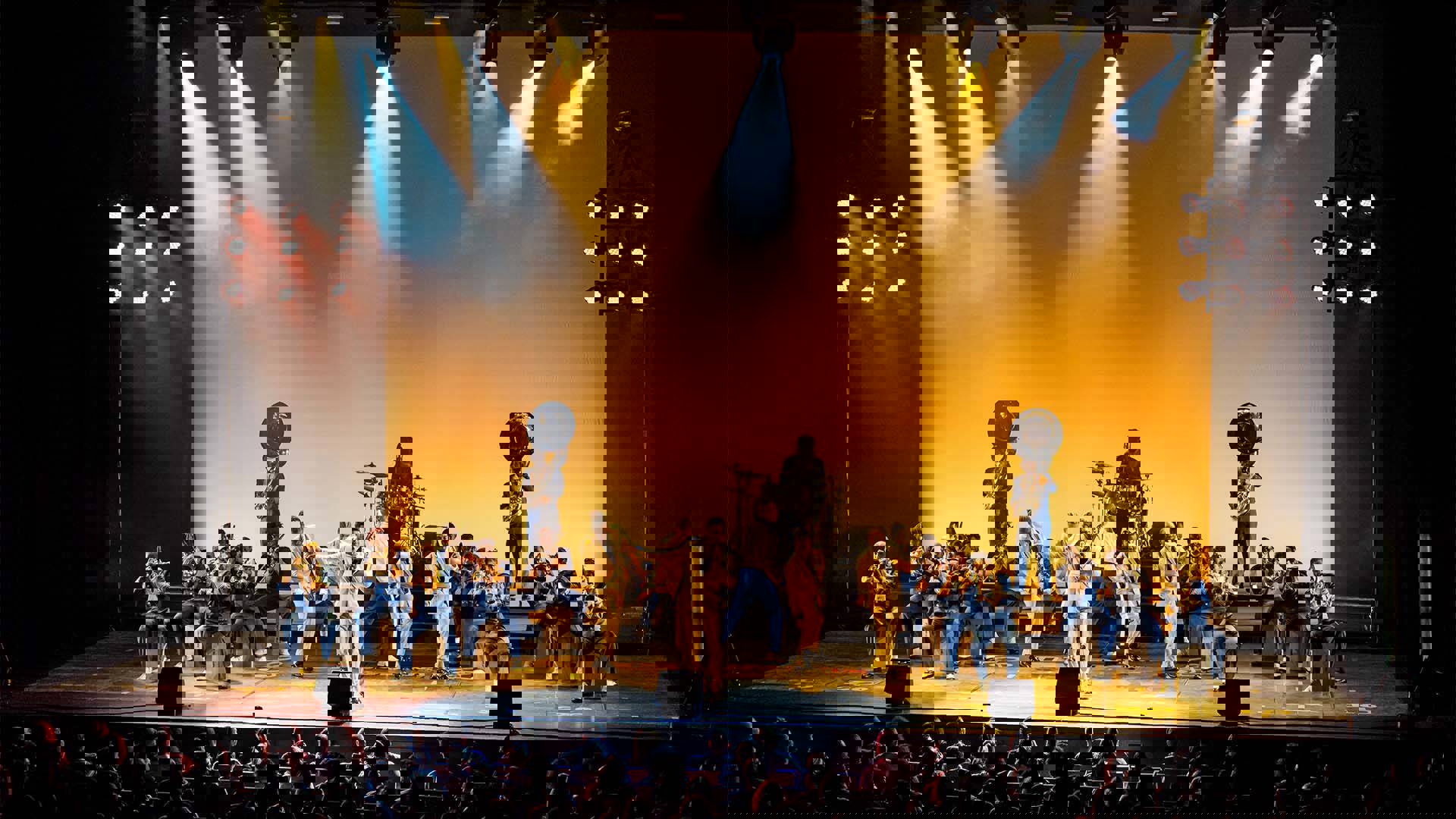 Drumline Live's African-American marching band performing on stage in blue uniforms. There are five performers in yellow suits singing and dancing center stage.
