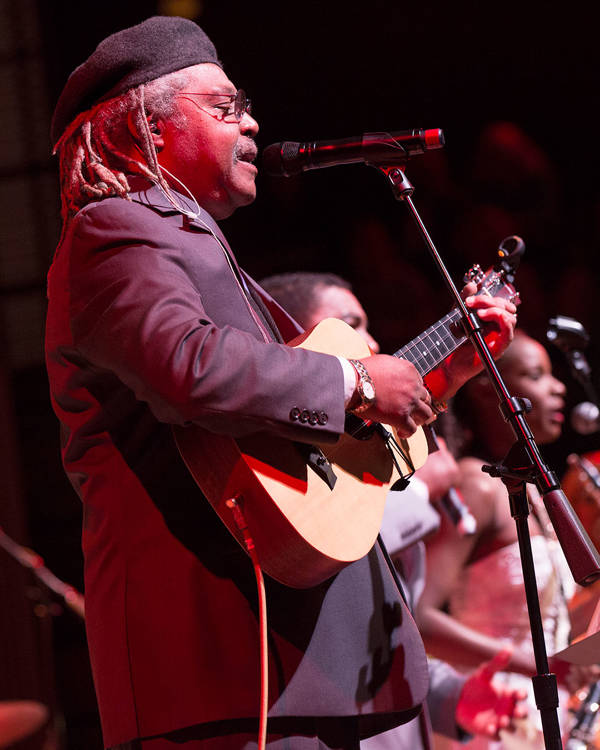 A man with dreadlocks, a black hat and a guitar is singing into a microphone.