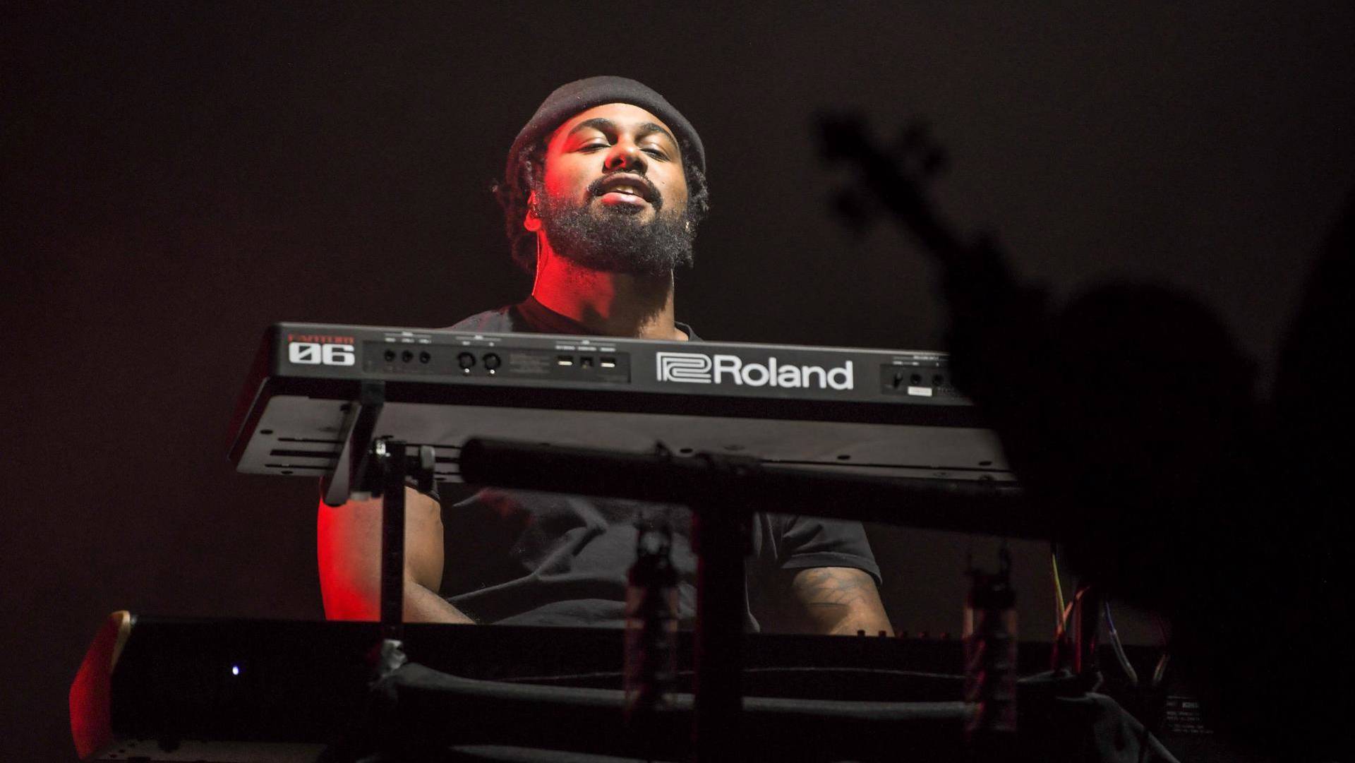 A man with dark complexion, a dark beard and dark clothing playing keyboards.