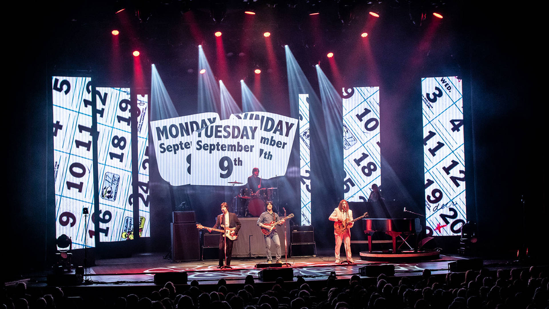 Four Beatles impersonators performing on a brightly lit stage, wearing stylish 1960s street clothes and holding guitars.. Various calendars and dates are projected behind them.