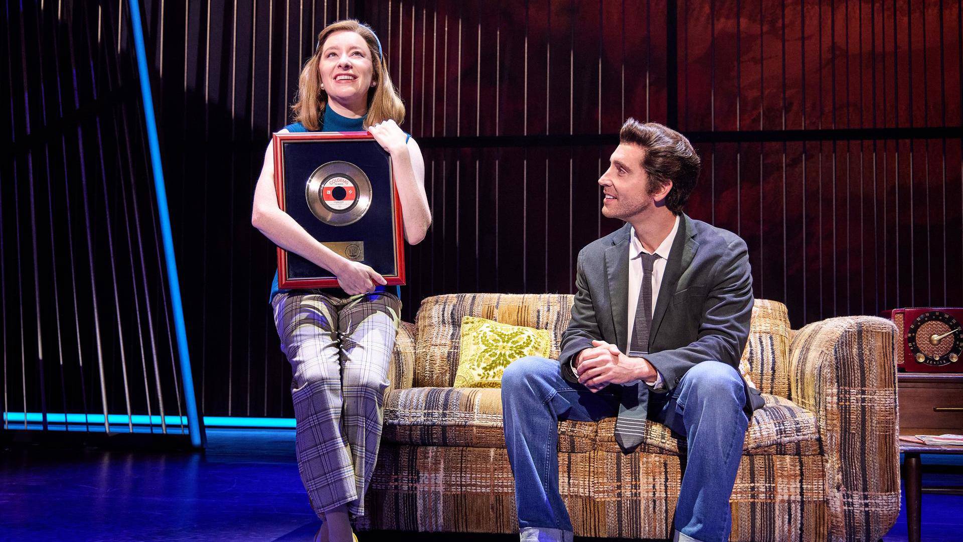 A man with light complexion and greased brown hair in a grey jacket and tie sits on an old couch and looks to his right at a woman with light complexion and medium blonde hair. She is holding a framed music award.