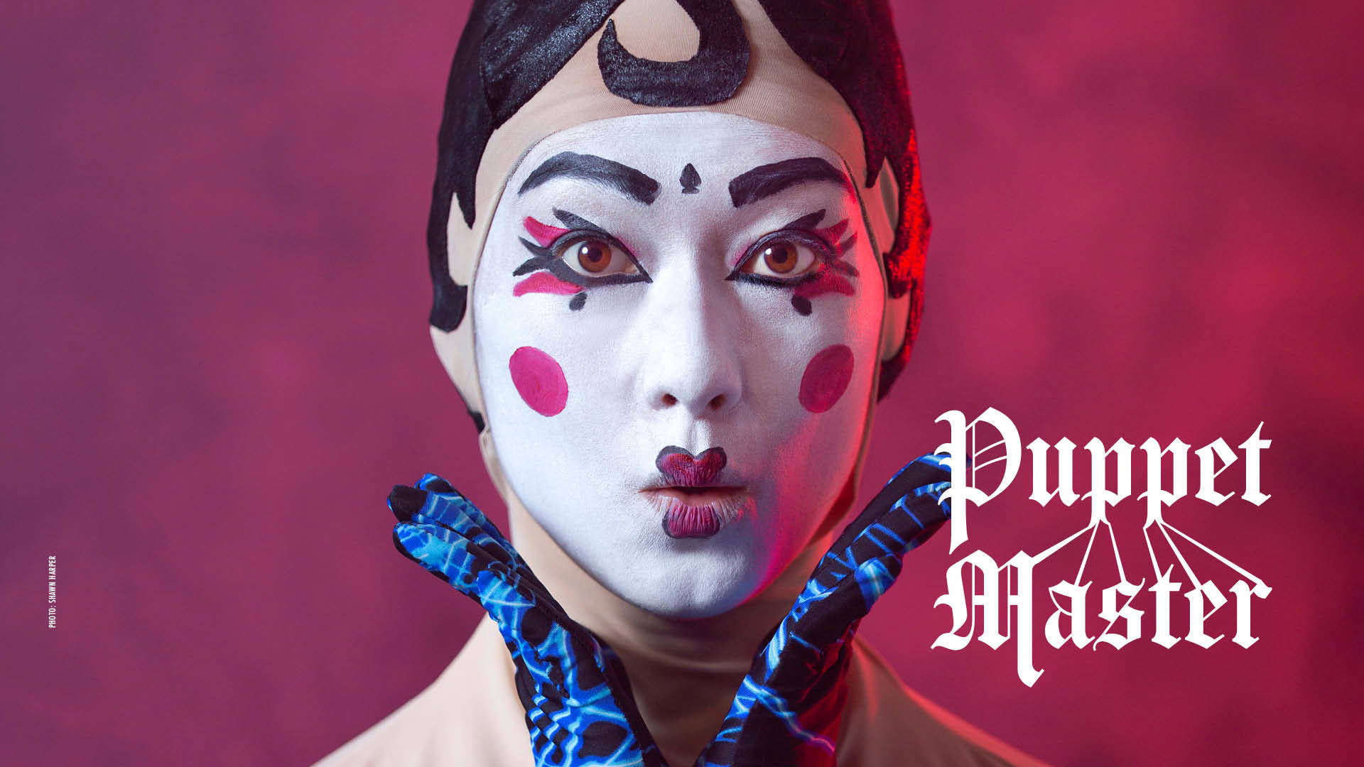 Banner reads: "Puppet Master." A close-up of a dancer's face against a red backdrop wearing white face paint with black-lined eyes and eyebrows, red circles for blush and heart-shaped red painted lips. Their lips are pursed and two blue-gloved hands frame the face.