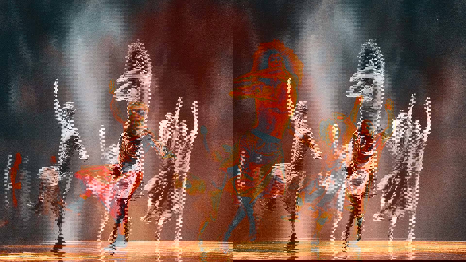 Five male dancers performing on stage in loose clothing, their arms are out wide and stepping to the side.