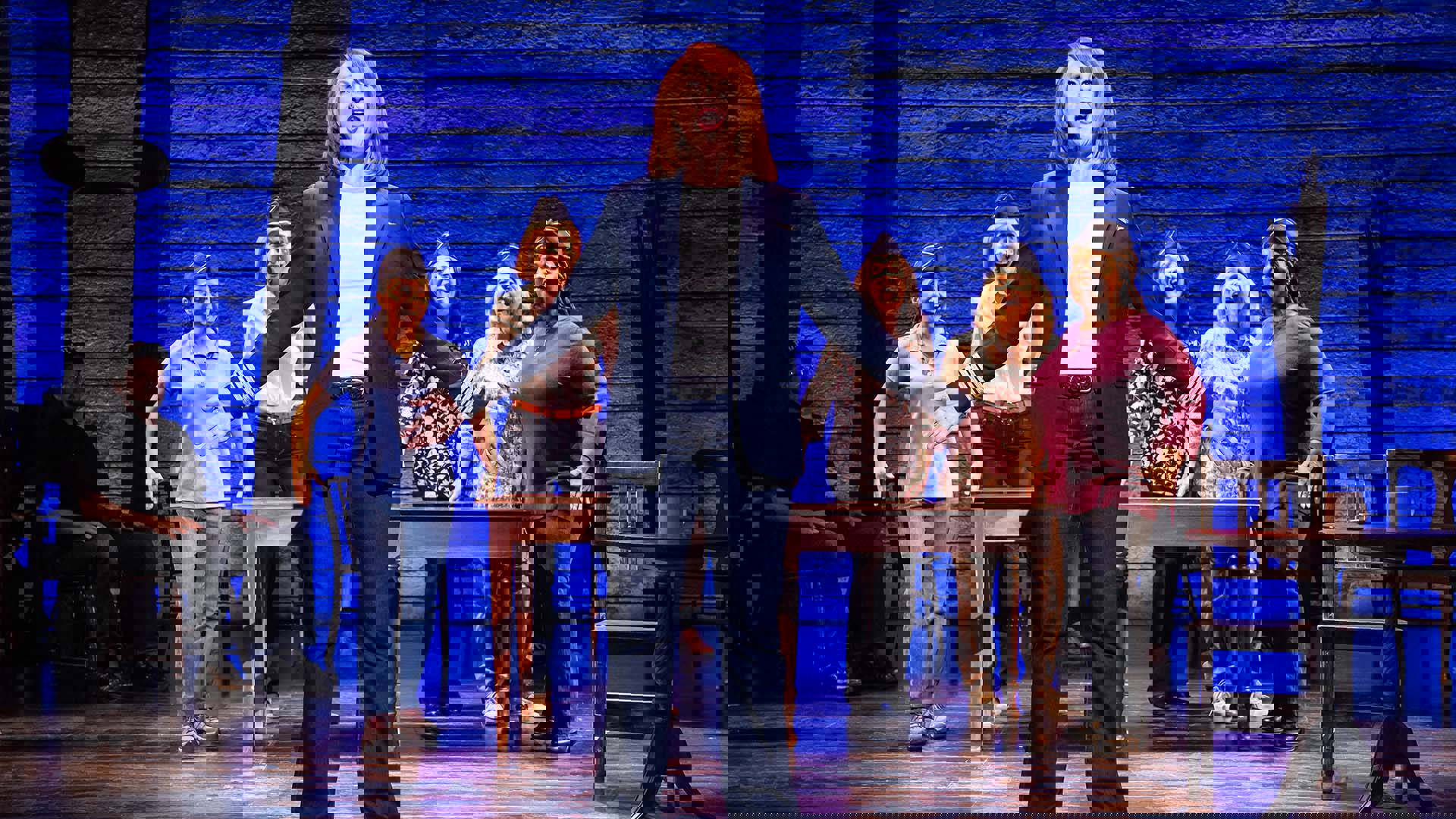 A white woman in a blue suit jacket and red hair sings in front of fellow cast members