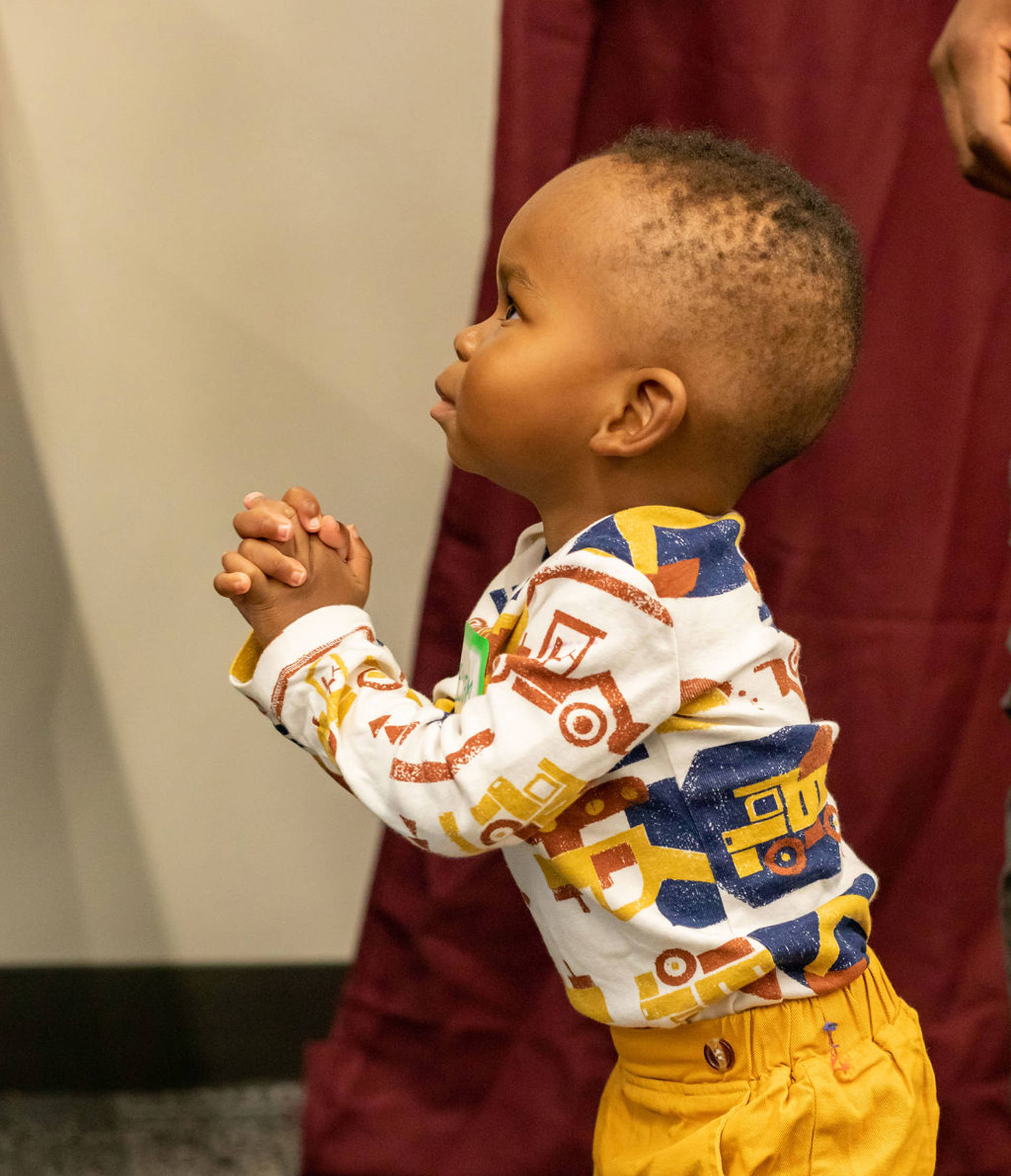 An toddler with dark complexion and brown eyes stands in profile to the viewer with his hands clasped.