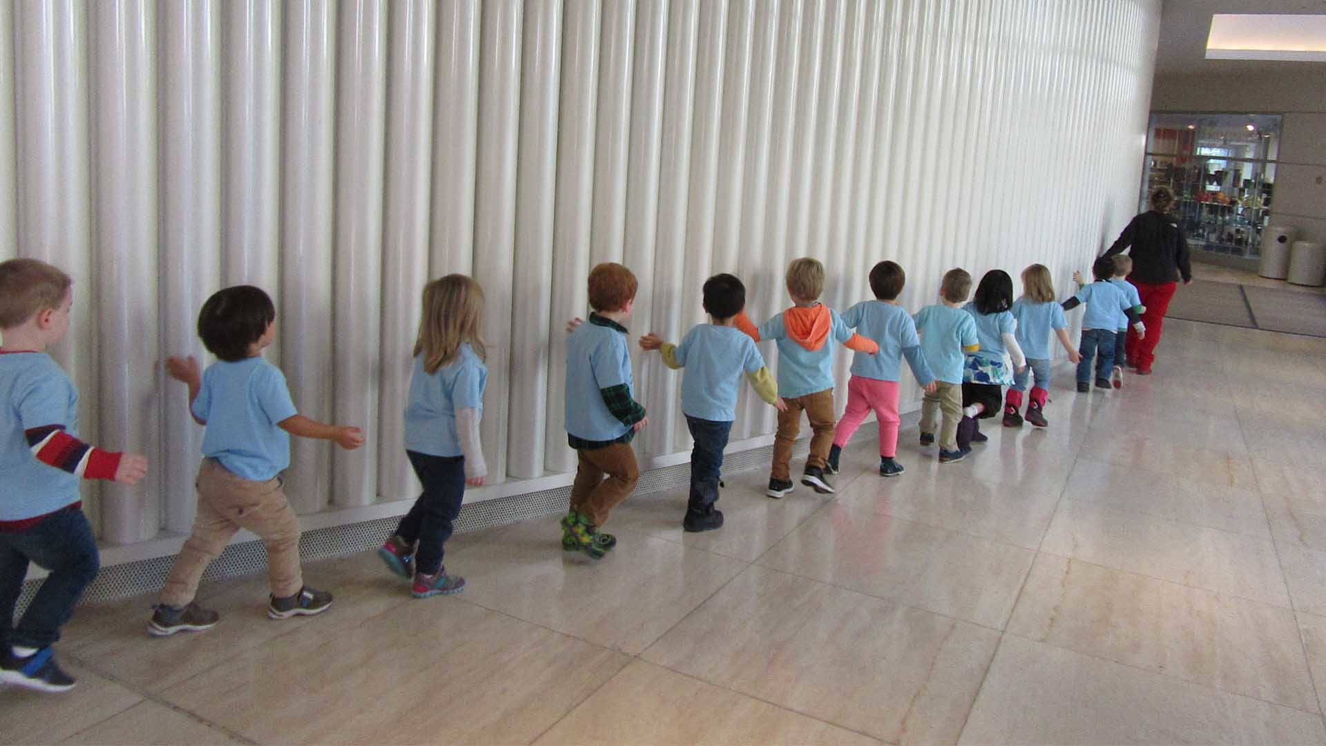 Young kids walking in line against a textured wall. They are touching the wall as the walk past it.