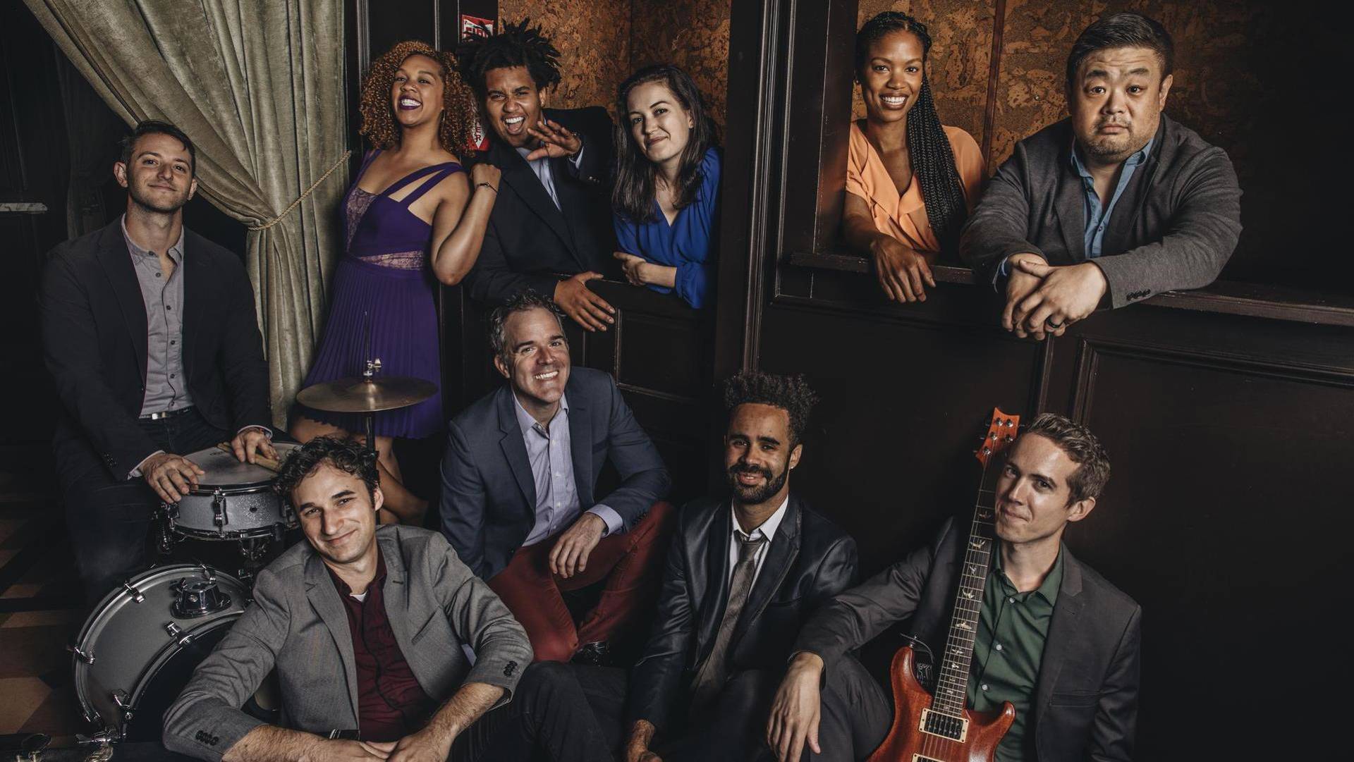 A diverse group of adults in semi-formal clothing smiling in a group photo in an ornate moody theater backdrop. One man is behind a drum kit and another is holding an electric guitar.