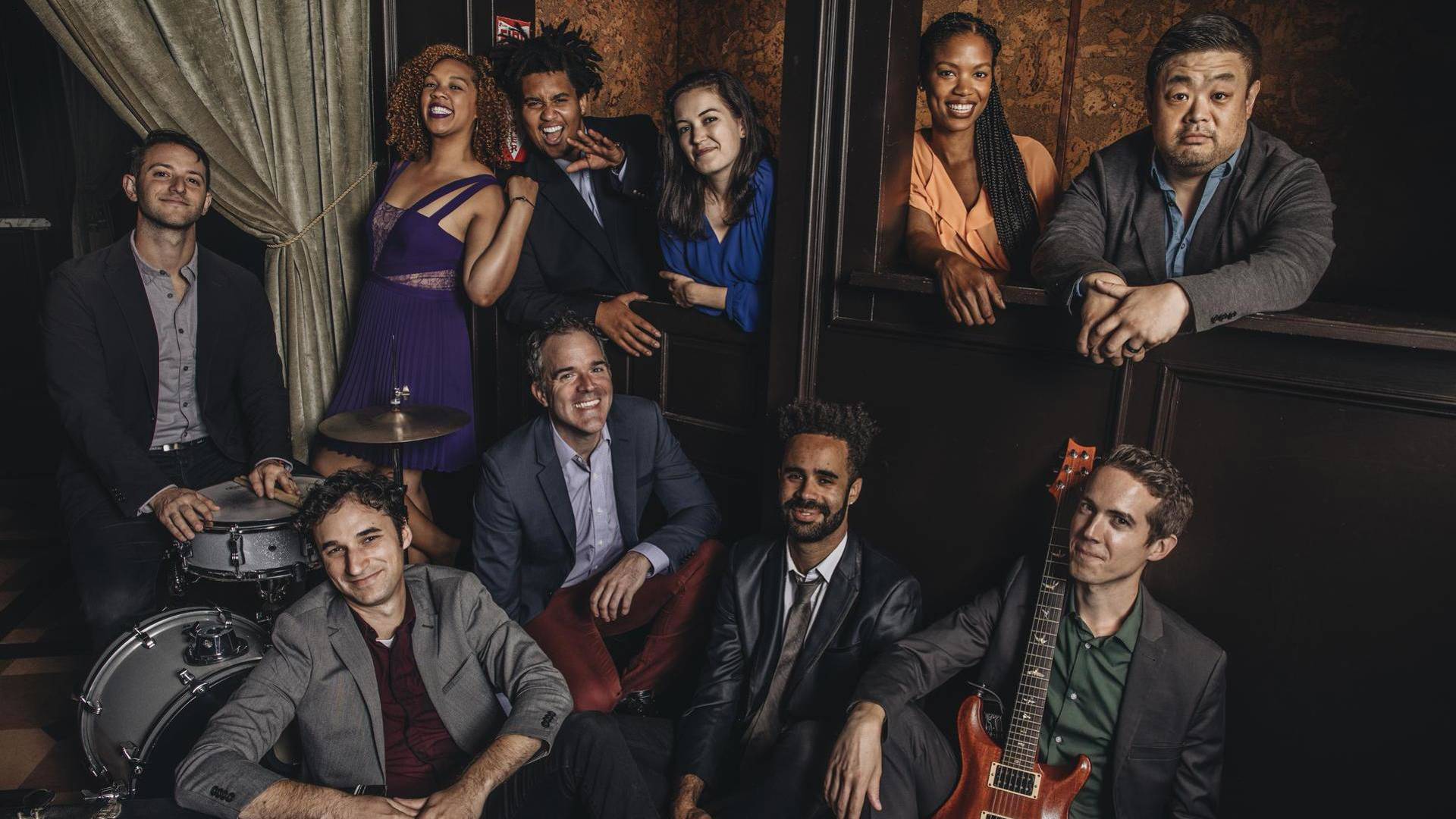 A diverse group of adults in semi-formal clothing smiling in a group photo in an ornate moody theater backdrop. One man is behind a drum kit and another is holding an electric guitar.