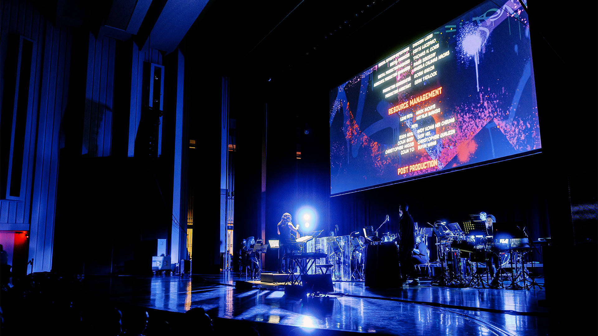 An orchestra on stage in dim blue light with spiderman into the spiderverse credits rolling on a large screen