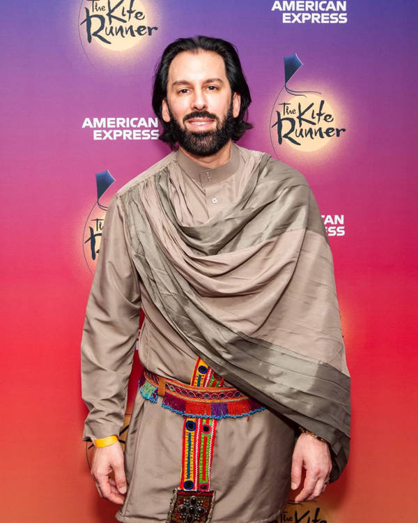 A man with medium complexion, medium dark hair and a dark beard stands and smiles. He is wearing a tan outfit, with a flowing component covering his left arm and a colorful piece around his midsection.