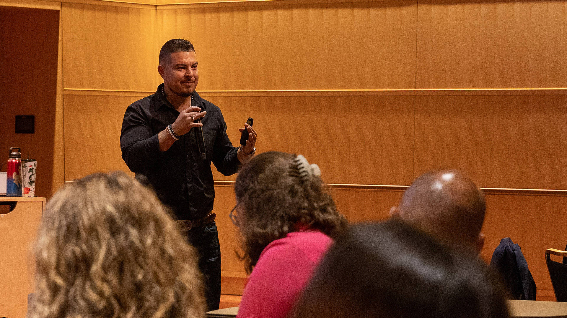A man with medium skin tone, dark short hair and a black dress shirt speaks to an audience.