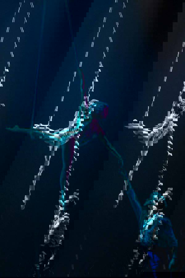 A woman with dark, short hair hangs acrobatically from the ceiling by a rope while holding the hand of a light-skinned man standing on the ground.