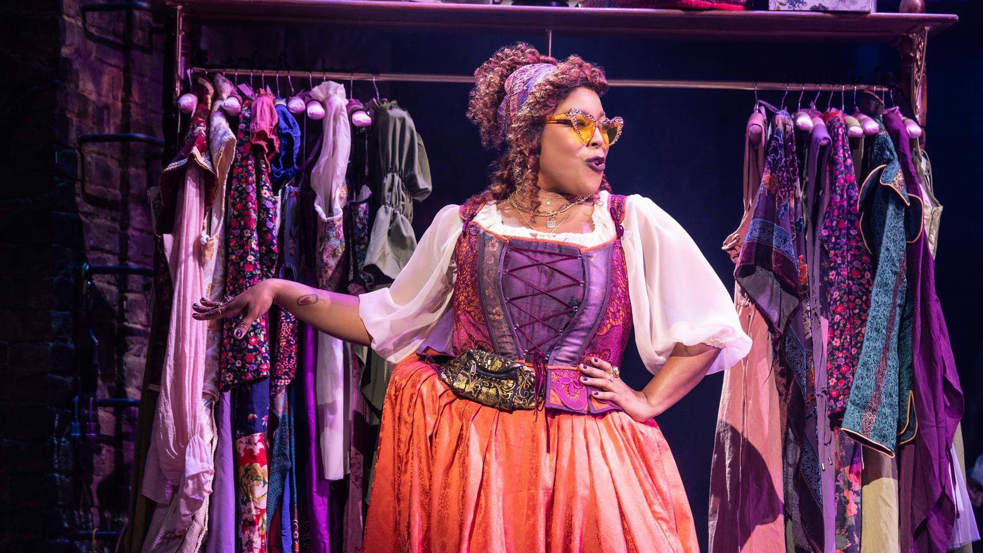 A woman with dark complexion, dark curly hair, a salmon and purple dress, and sunglass sings in front of a rack of colorful clothing.