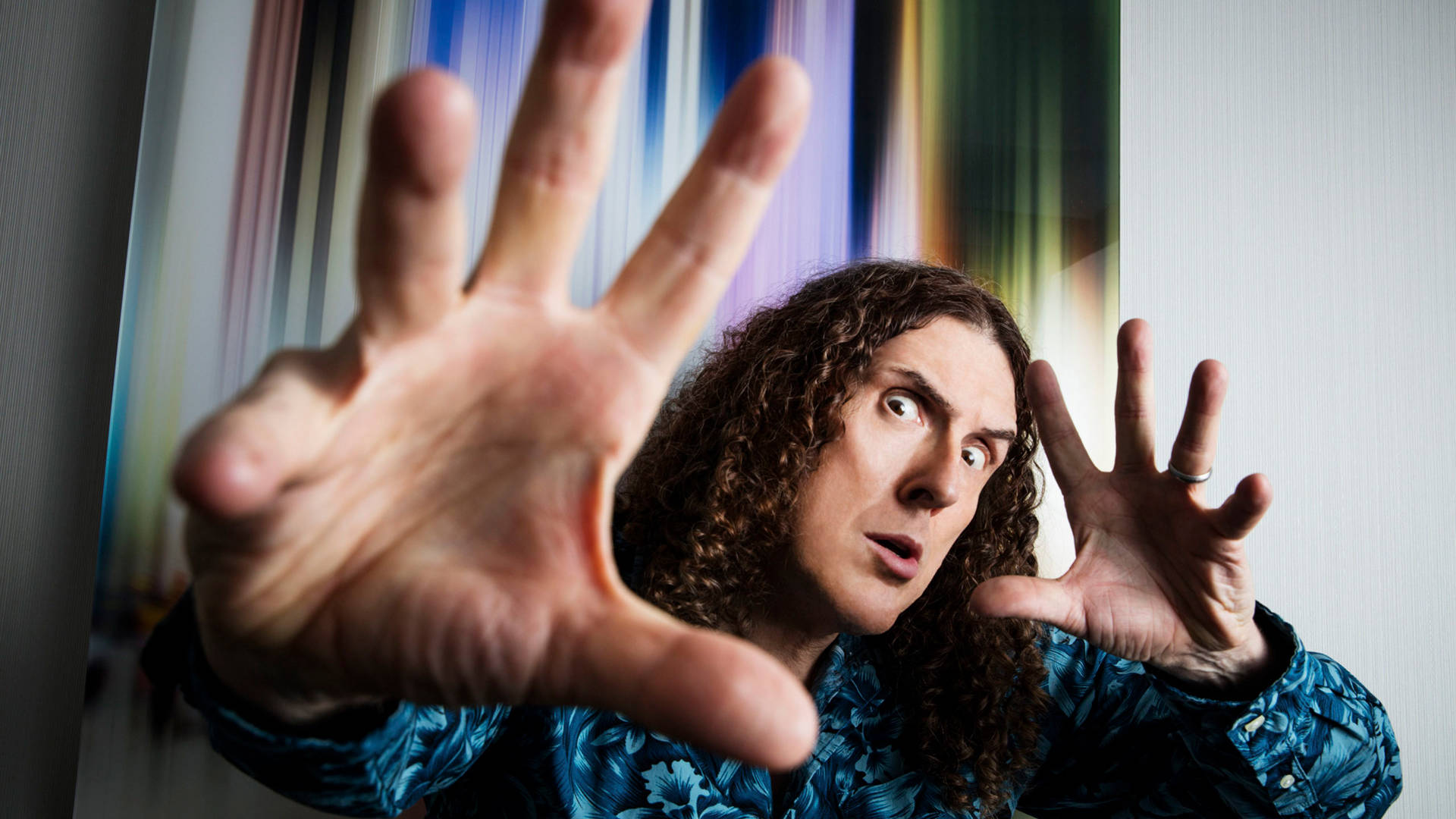 "Weird Al" Yankovic is looking at the viewer and holding out his hands as if he is casting a spell. He has a blue shirt and iconic curly hair