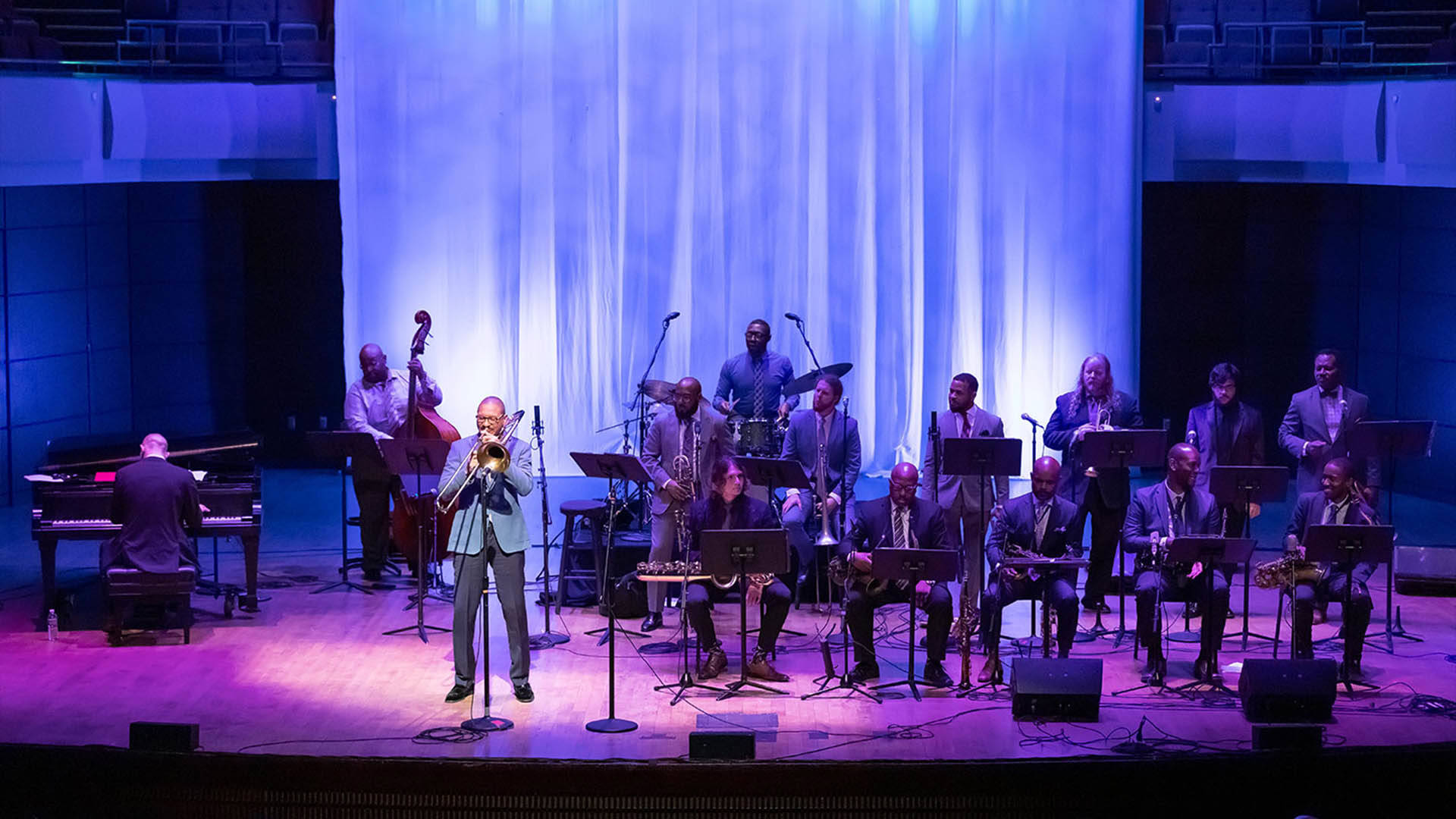 A jazz band of 15 members performing on a stage. A solo trombonist stands in a spotlight. The stage has blue and purple lighting and a large curtain that looks like a waterfall hangs behind the band.