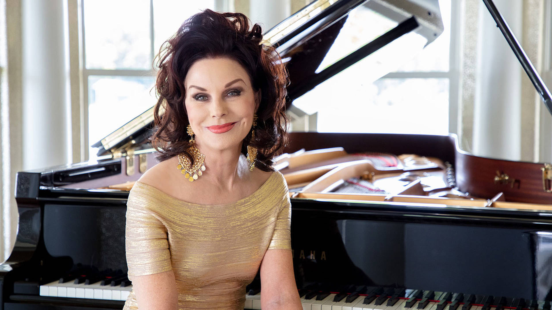 A woman with light complexion, dark hair, and a gold dress with gold earings posing for a photo. She is sitting in front of a piano.