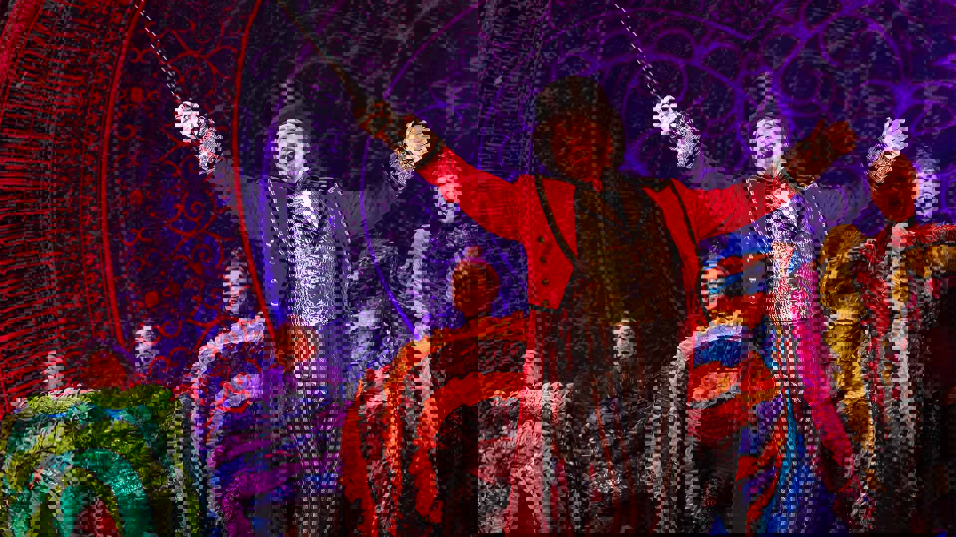 The cast of Moulin Rouge! in various colorful costumes performing together. A man in a red jacket, gold vest and tophat is at the center with his arms raised.
