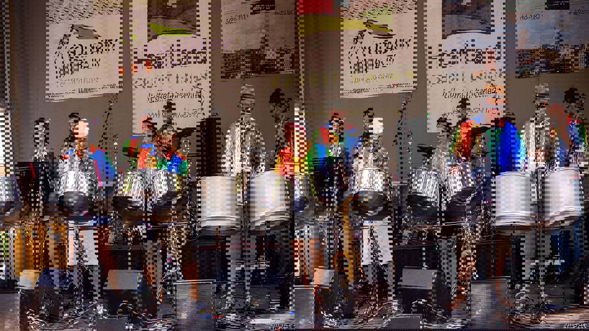 Seven adults in rainbow tie-dye shirts performing large steel drums on an outdoor stage.