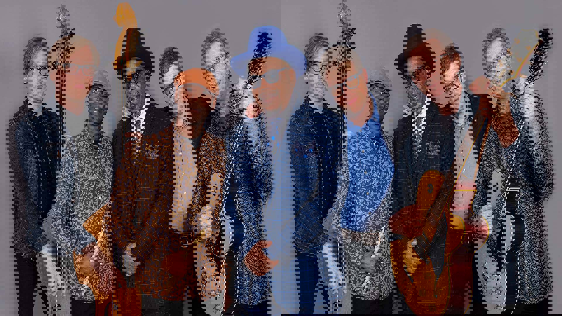A quintet of men stand in front of a grey backdrop. They are wearing an assortment of colorful shirts, jackets, and hats. The man on the left has an upright bass and the man on the left has a guitar.