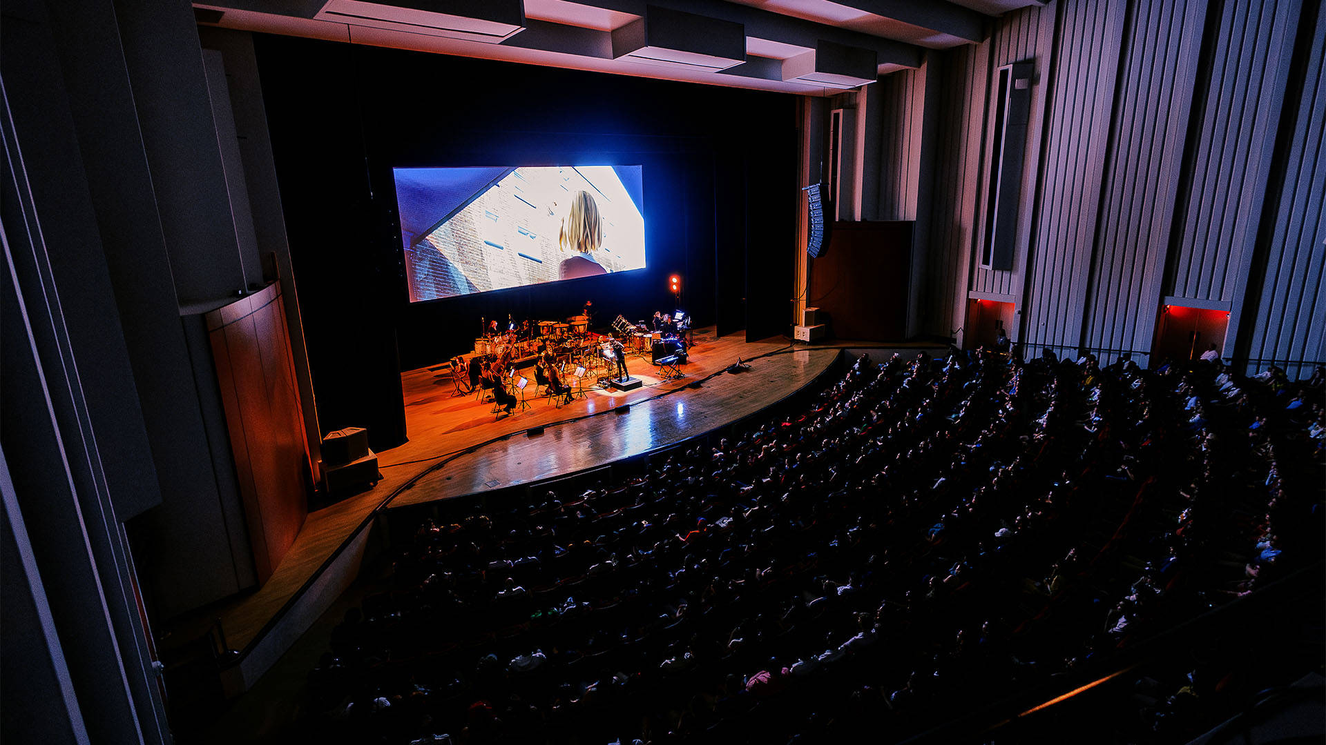 A full house audience watching an orchestra performing on stage under a large screen with a scene from Spiderman into the Spider-verse.