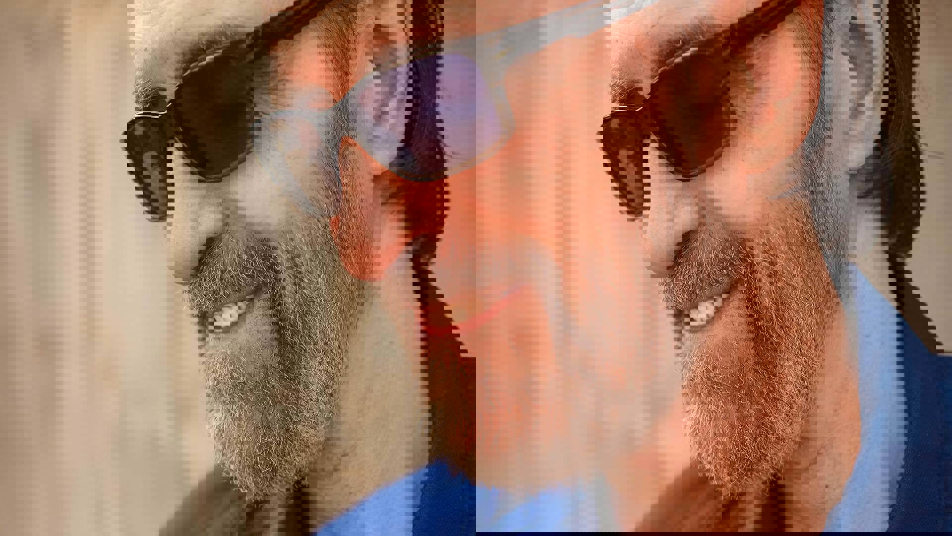 Close up of a smiling George Winston. He has sunglasses on, and a short brown and grey beard and mustache.