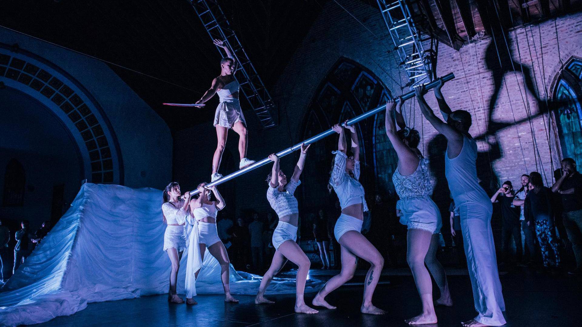 Six acrobats with light skin tones in white clothing stand while holding a large metal beam on stage. Another acrobat balances on the metal beam with their hands raised for balance.