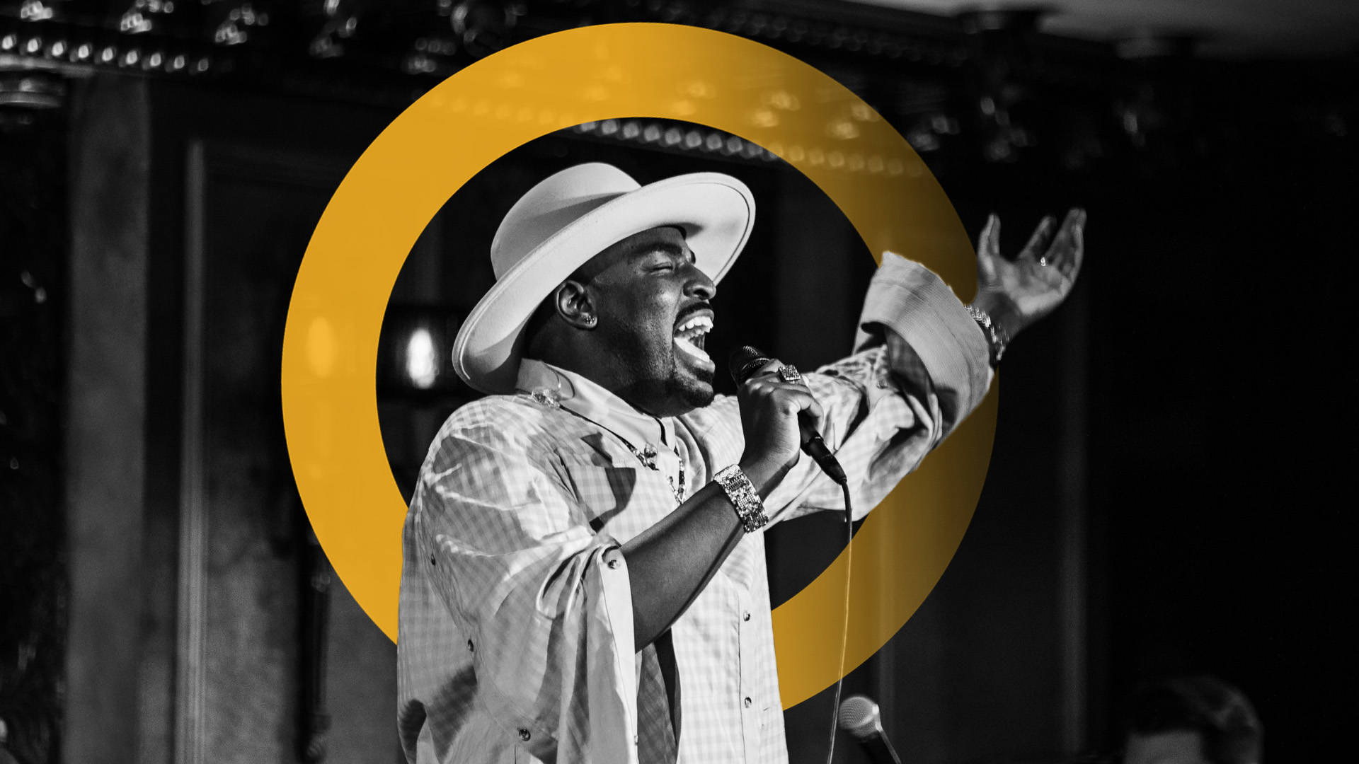 A black man wearing a plaid collared shirt with the sleeves rolled up to the elbow. He has a light colored wide-brimmed hat on and is singing passionately into a microphone on stage with one arm out wide.