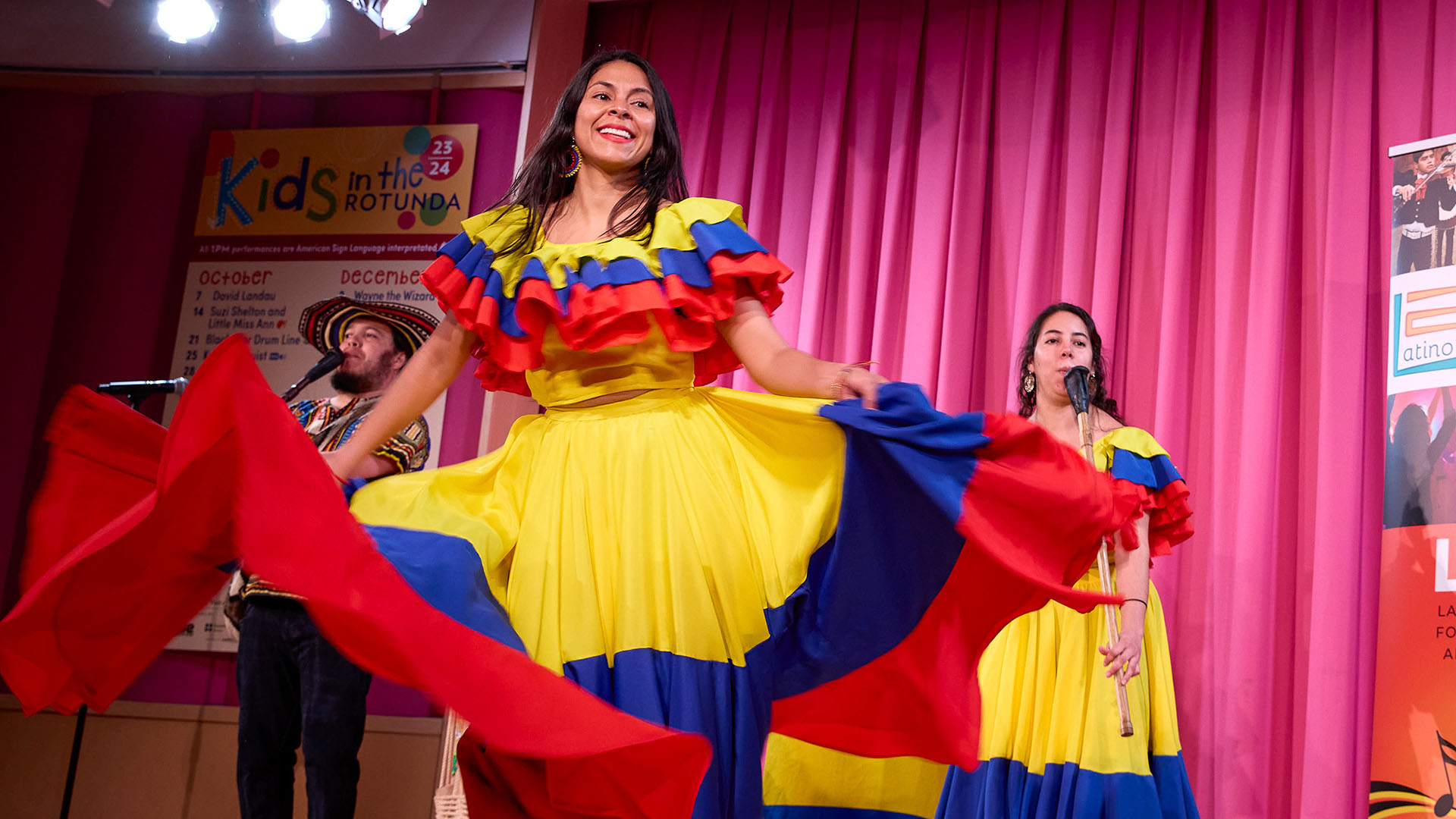 A woman with medium complexion twirls in a yellow, blue and red traditional latin dress on Rotunda Stage. She is smiling with long dark hair and hoop earrings.