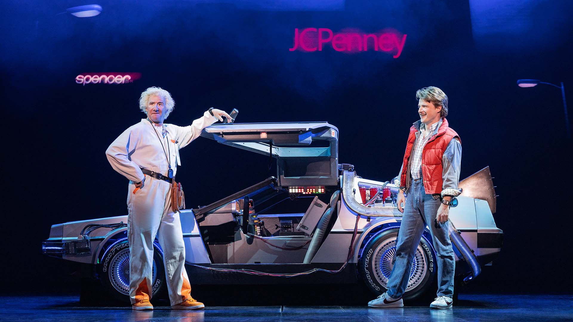 An older man with wavy white hair and a light complexion speaks to a young man with brown hair and a light complexion in front of a Delorian.