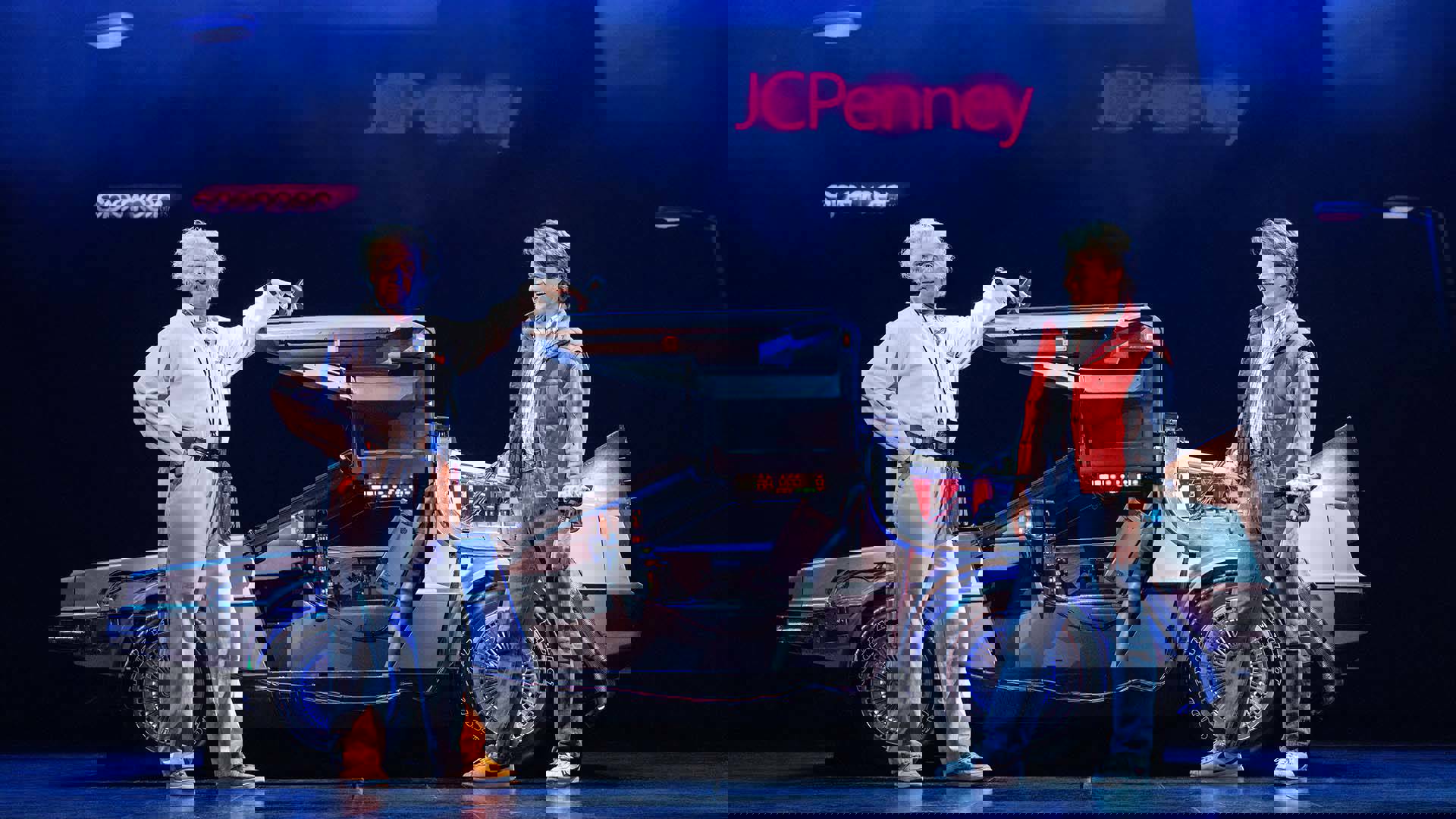 An older man with wavy white hair and a light complexion speaks to a young man with brown hair and a light complexion in front of a Delorian.