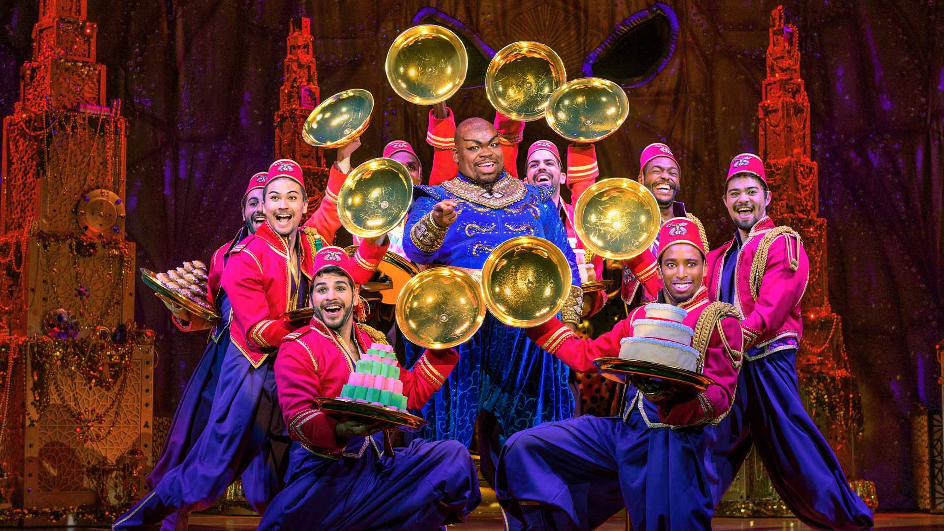 A Black man in a purple, robed outfit pointing at the camera. He is surrounded by eight other men wearing pink fezzes, pink jackets and purple pants. The men hold various deserts on trays. In their other hands they hold gold tray covers in a circle around the purple robes.