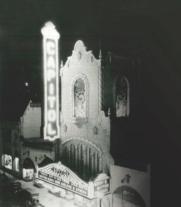 A black and white photo of the Capitol Theater marquee. It stands vertically and lit up brightly at night.