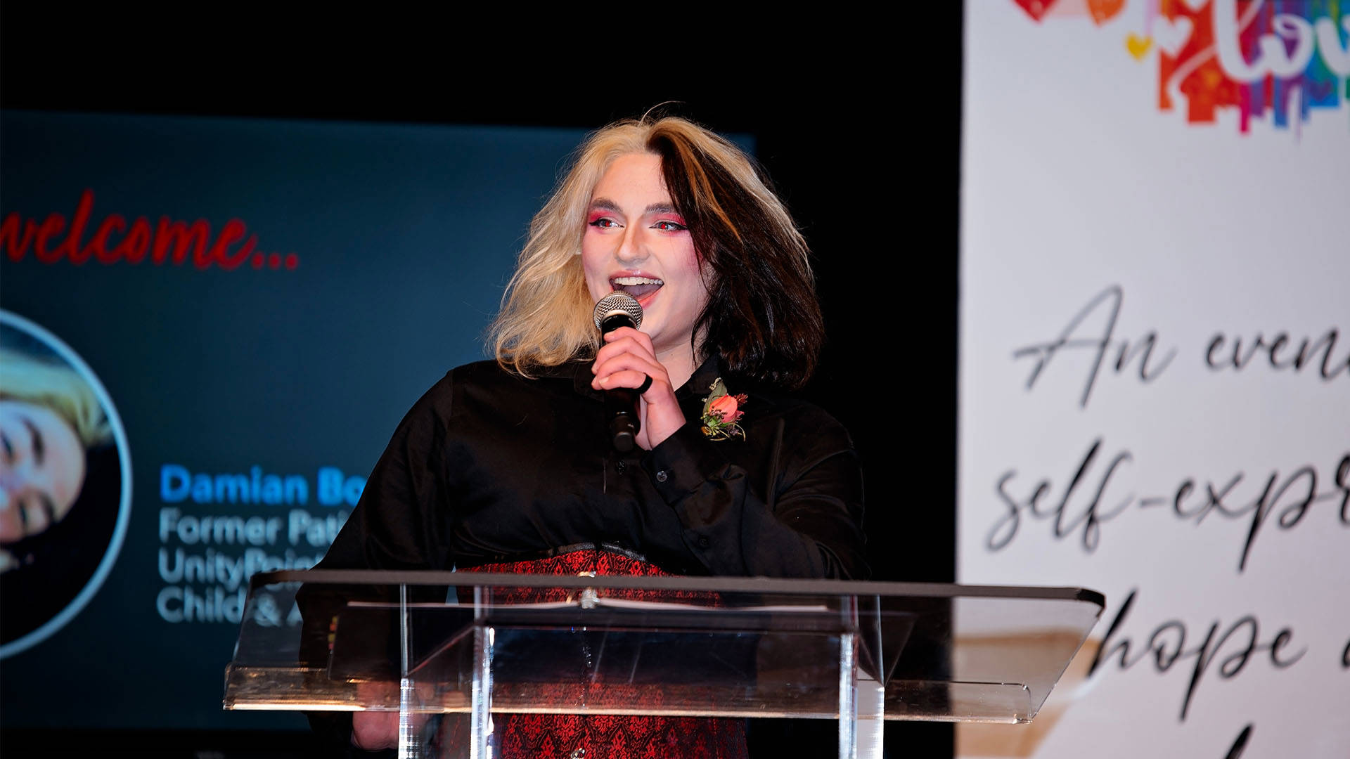 A person with standout eye shadow and dark hair highlighted blond speaking at a podium