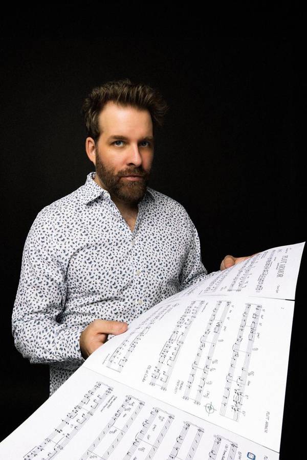 A young man with medium complexion, short brown hair, a beard, and a light shirt. He is holding a piano score.