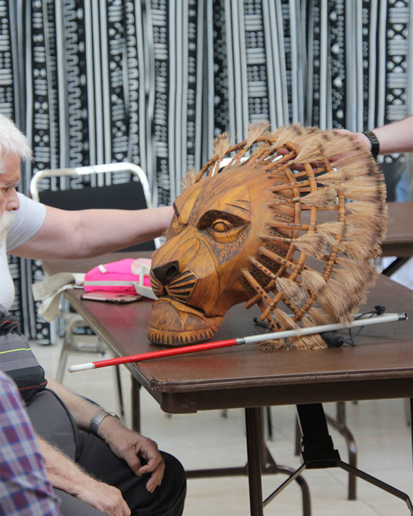 A wooden lion mask on a table. Outstretched hands are touching it.