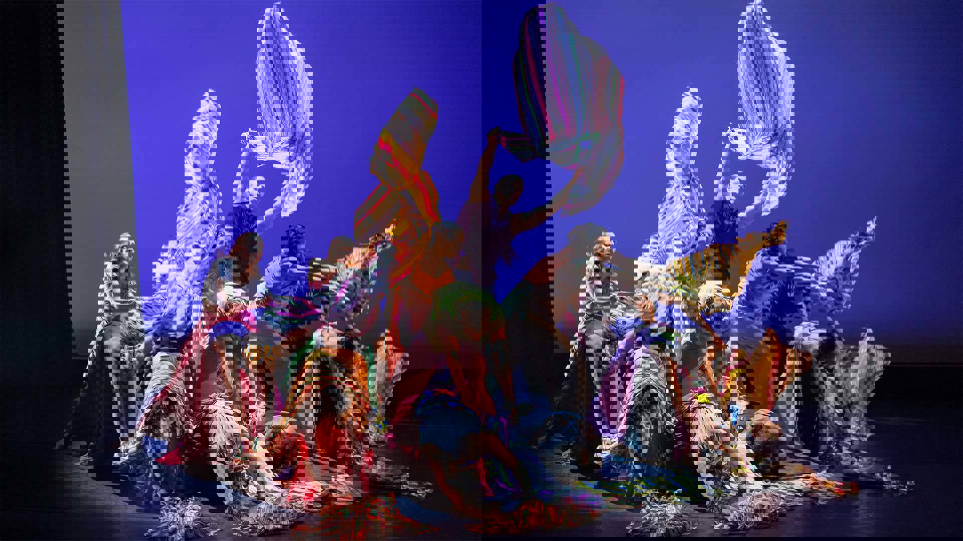 A group of a dozen dancers piled onto each other and draped in striped multicolor fabrics. Some are kneeling or standing and drifting the fabric above them.